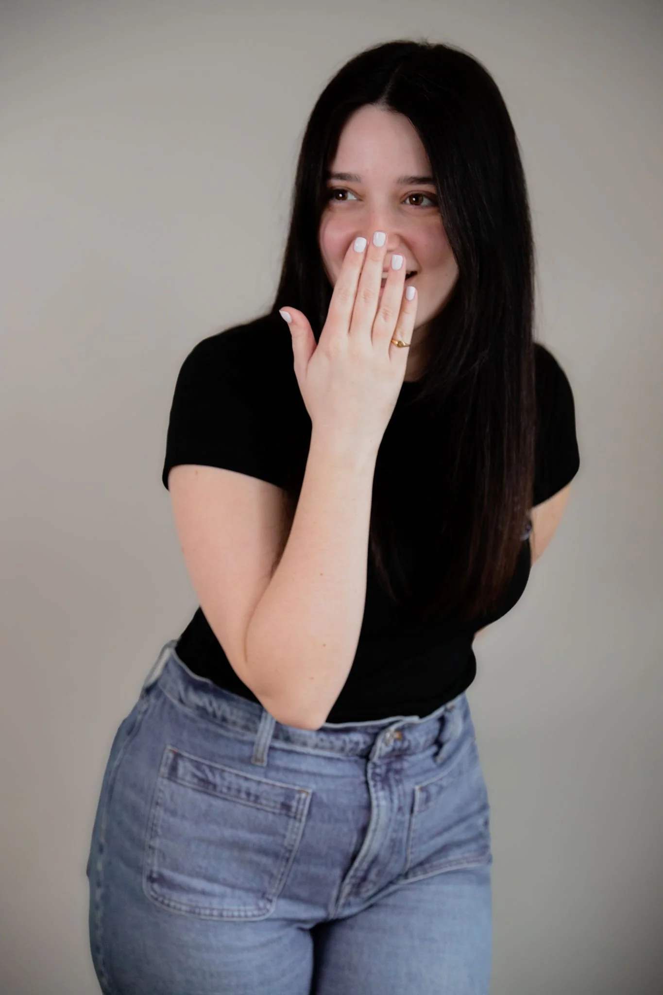A woman with long dark hair, wearing a black t-shirt and light blue jeans, covering her mouth with her right hand and smiling.