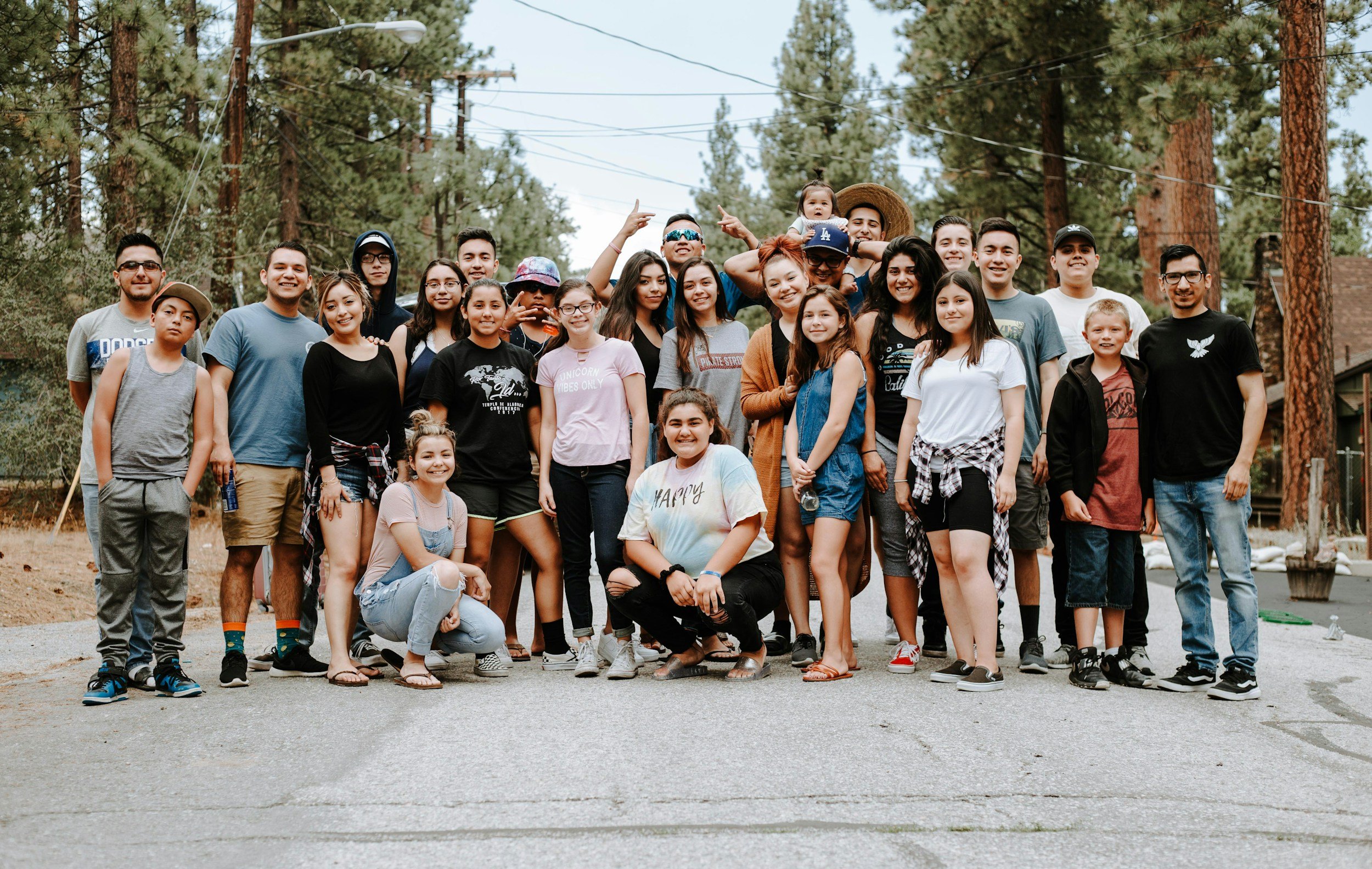A large group of diverse young people and children smiling outdoors in a wooded area with tall trees, power lines, and a cloudy sky, gathered together for a group photo.