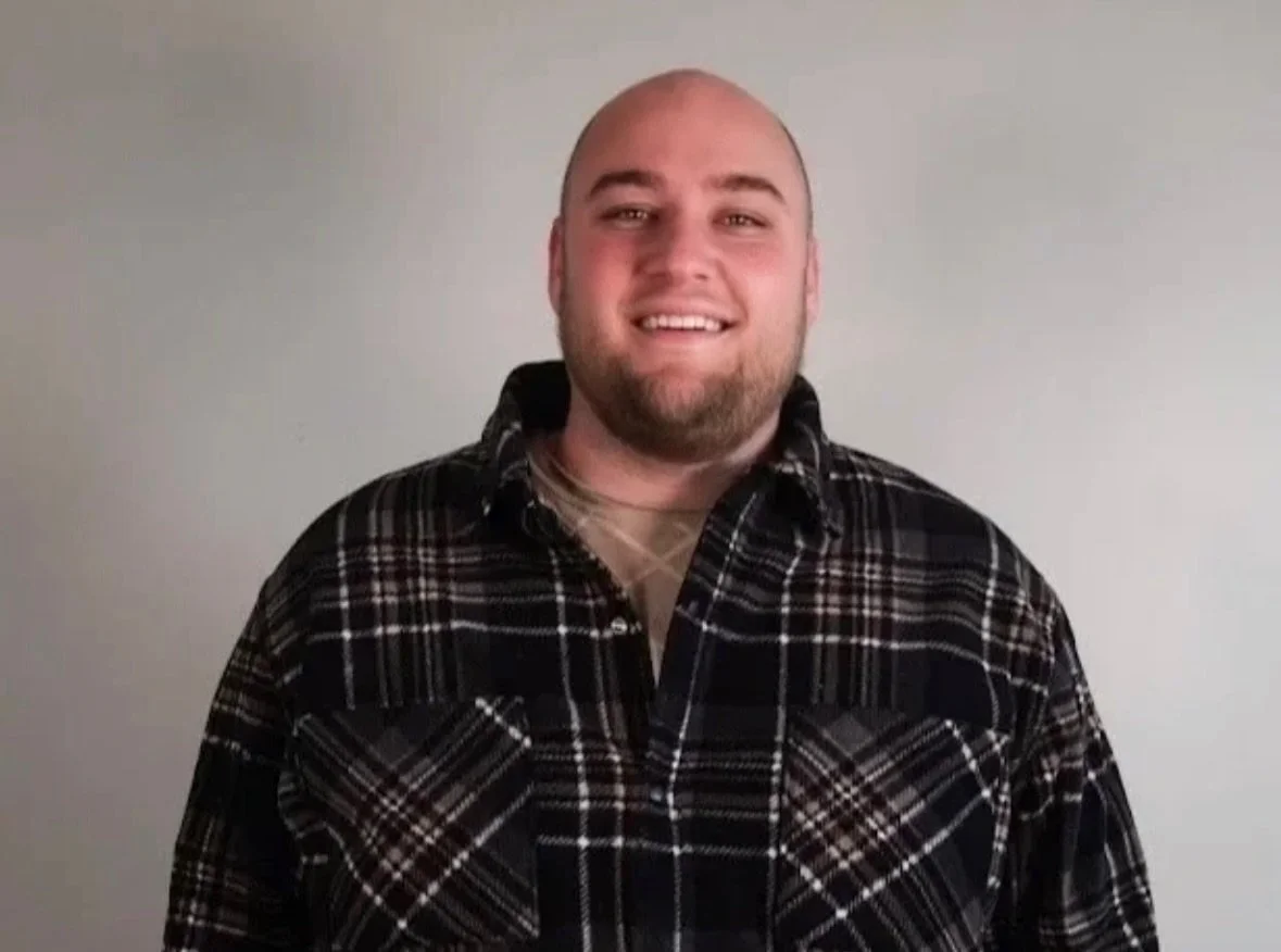 A smiling man with a beard, wearing a black and gray plaid shirt, standing against a plain light gray background.