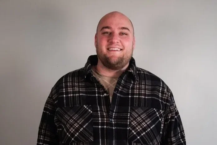 A smiling man with a beard, wearing a black and gray plaid shirt, standing against a plain light gray background.