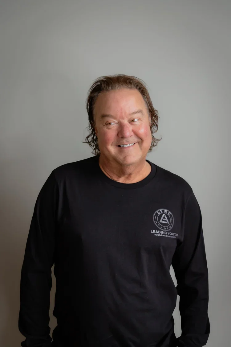 A middle-aged man with brown, wavy hair smiling and looking to the right, wearing a black long-sleeve shirt with a logo that reads "Leading Youth" and a geometric symbol.
