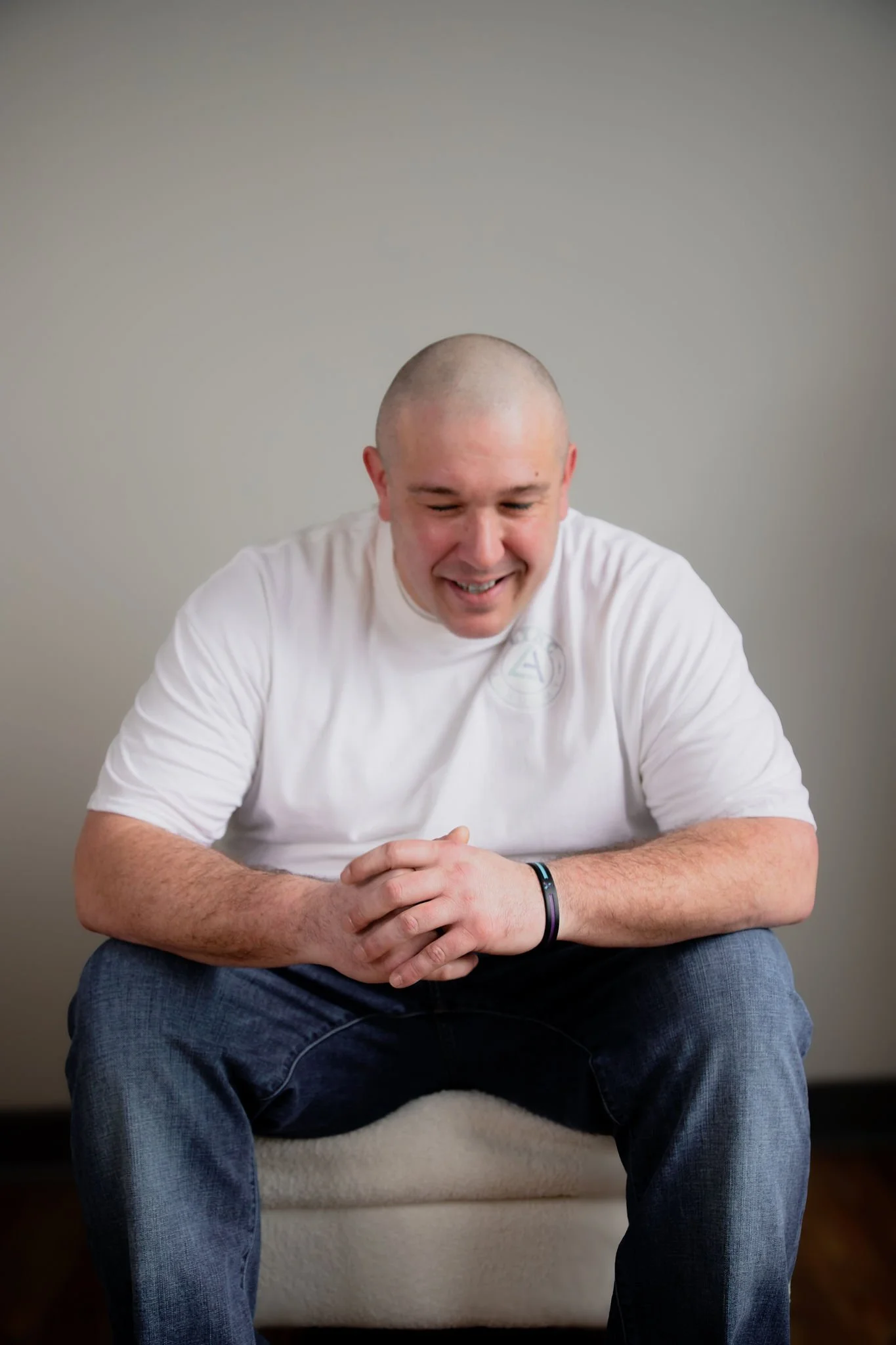 A young man with a shaved head sitting on a chair, smiling and looking down, wearing a white t-shirt and jeans.
