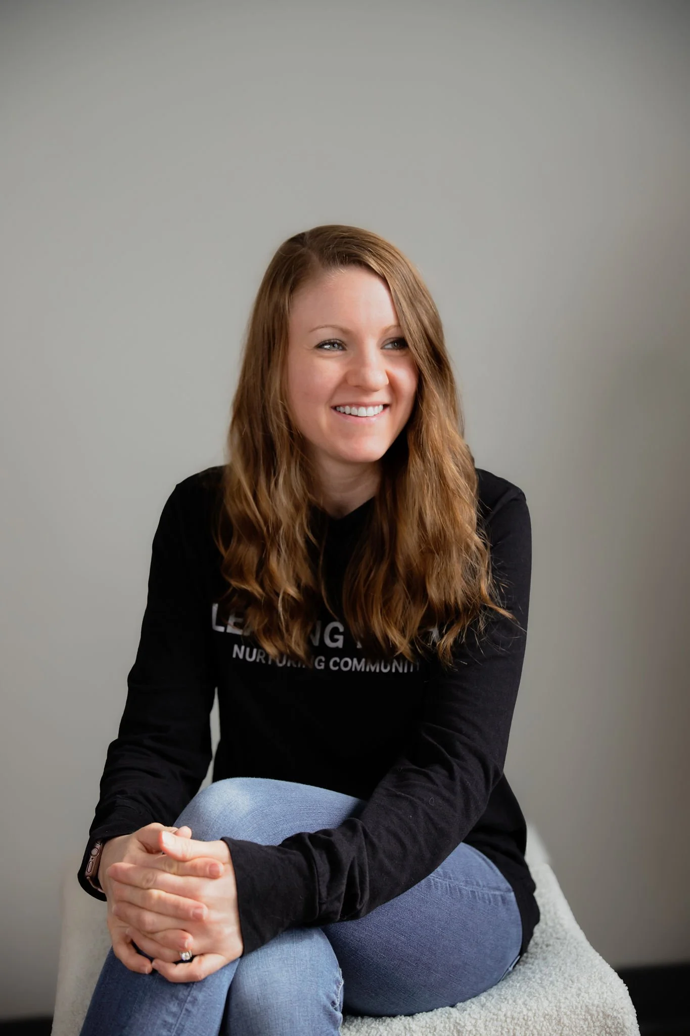 A woman with long wavy brown hair smiling and sitting on a cushioned stool against a plain gray background.