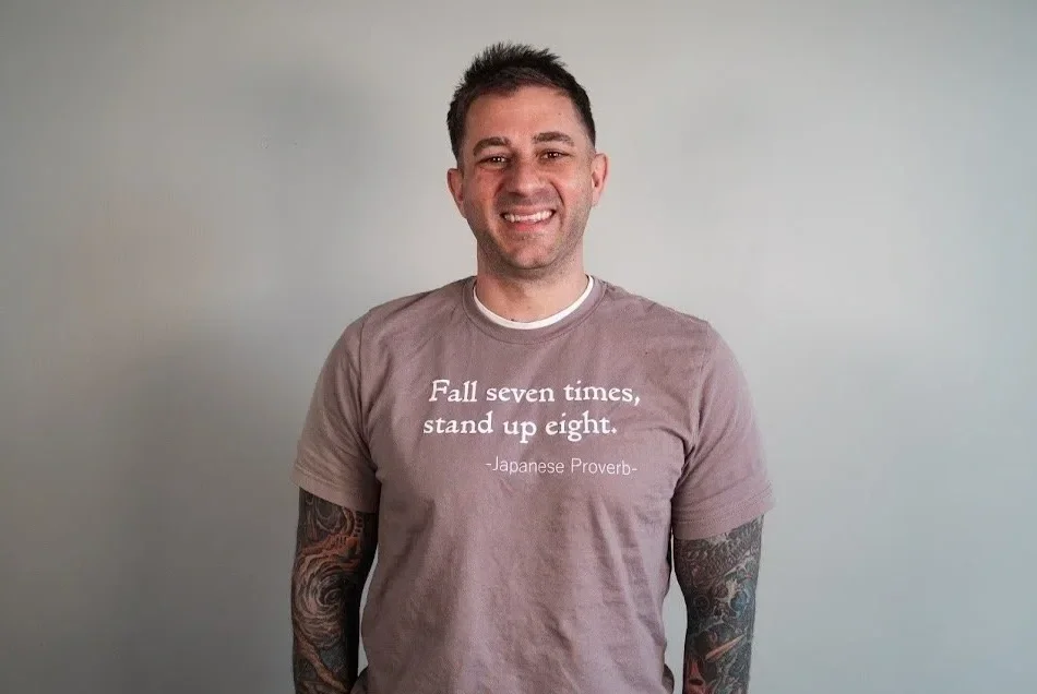 Smiling man with tattoos wearing a beige T-shirt with a Japanese proverb on it, standing against a plain light gray wall.