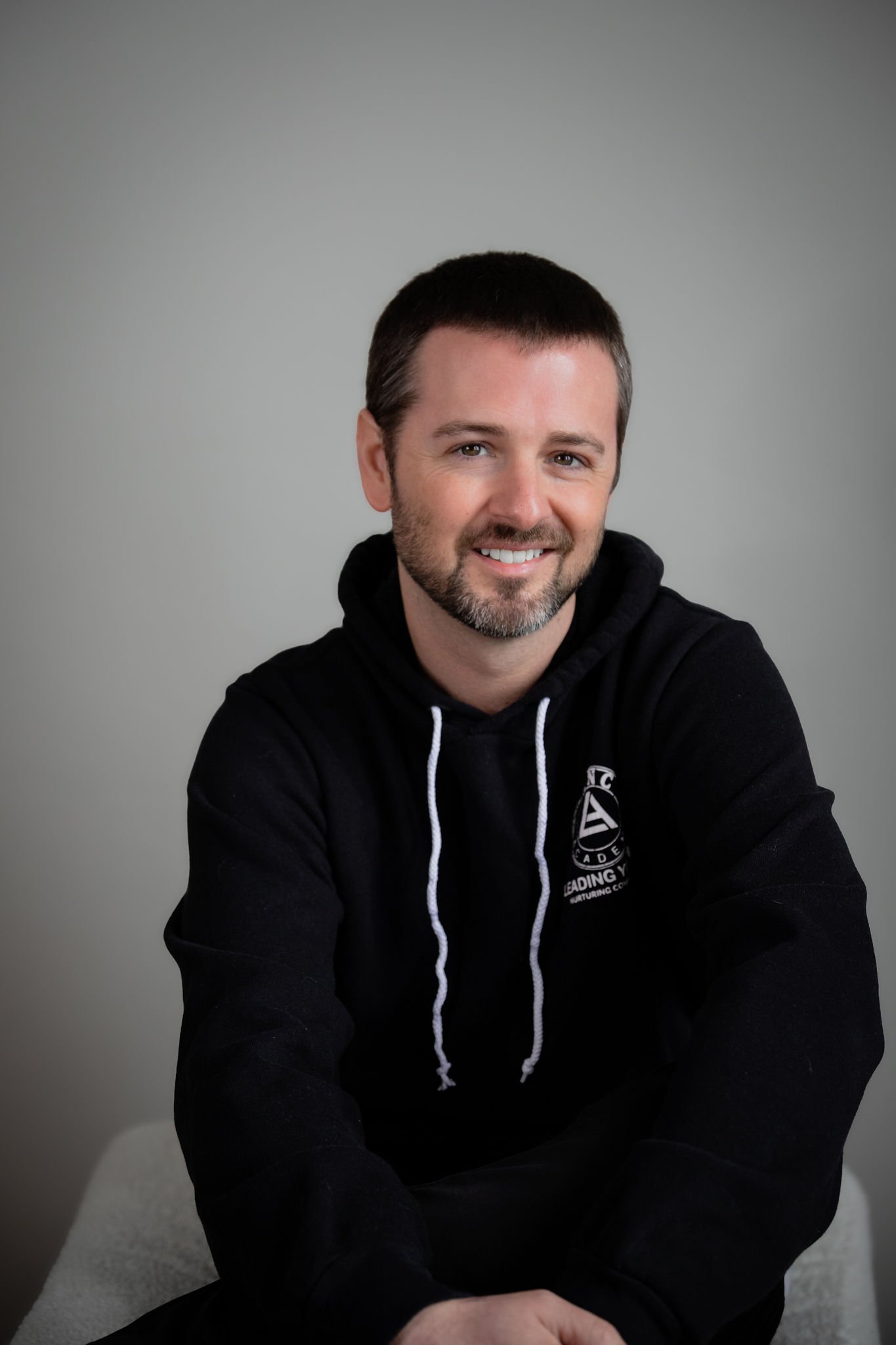 A man with short dark hair and a beard is smiling at the camera, wearing a black hoodie with white drawstrings and a logo on the chest, sitting against a plain gray background.