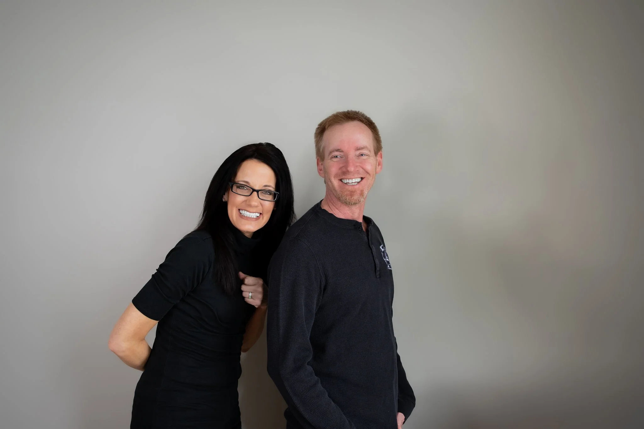 A woman with long black hair, glasses, and a black shirt next to a man with short light brown hair, a beard, and a black shirt, both smiling against a plain grey background.