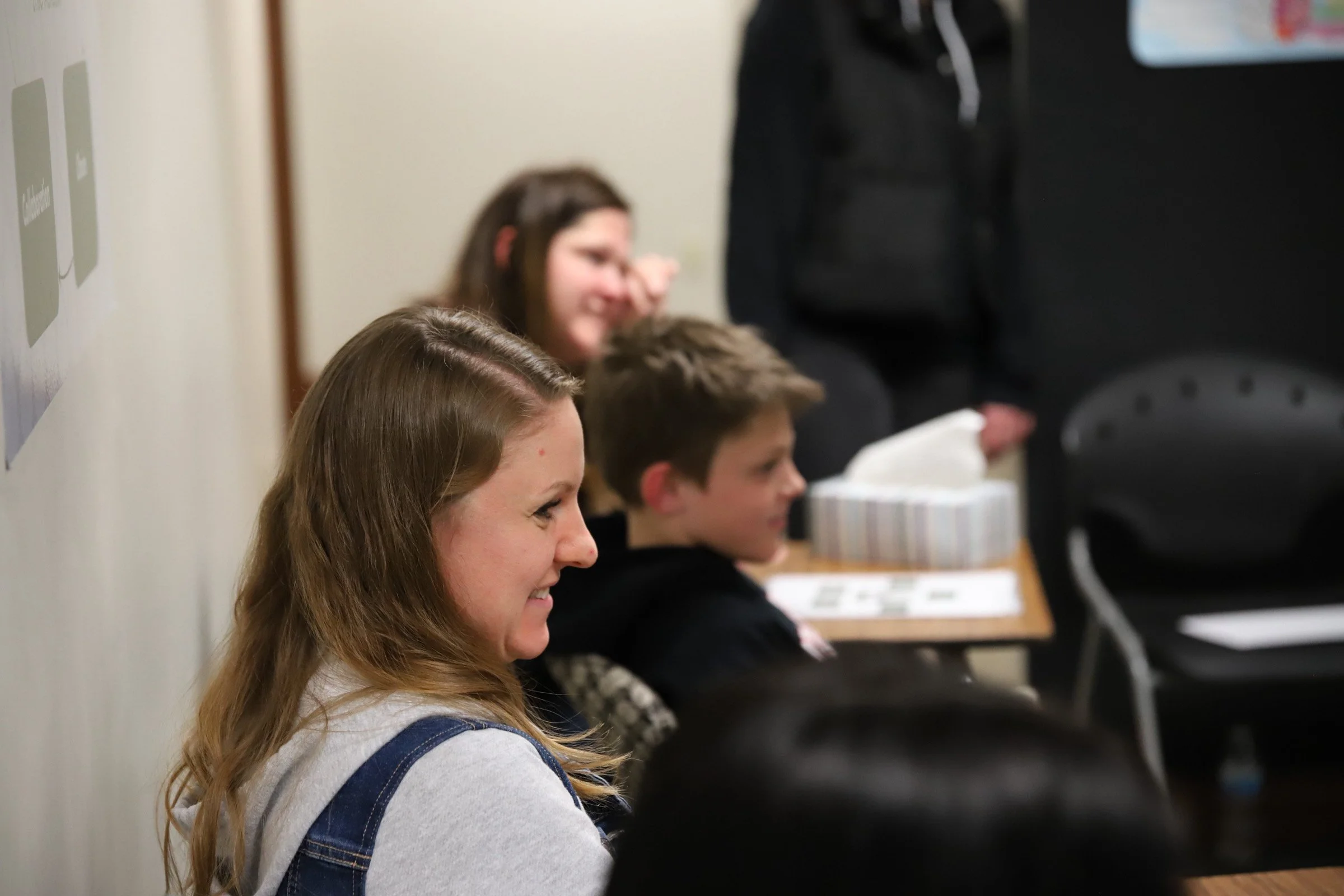 A group of four people sitting at a table, smiling and appearing to watch something off-camera. The group includes two women and two boys, with the woman in the foreground and the others slightly blurred in the background.