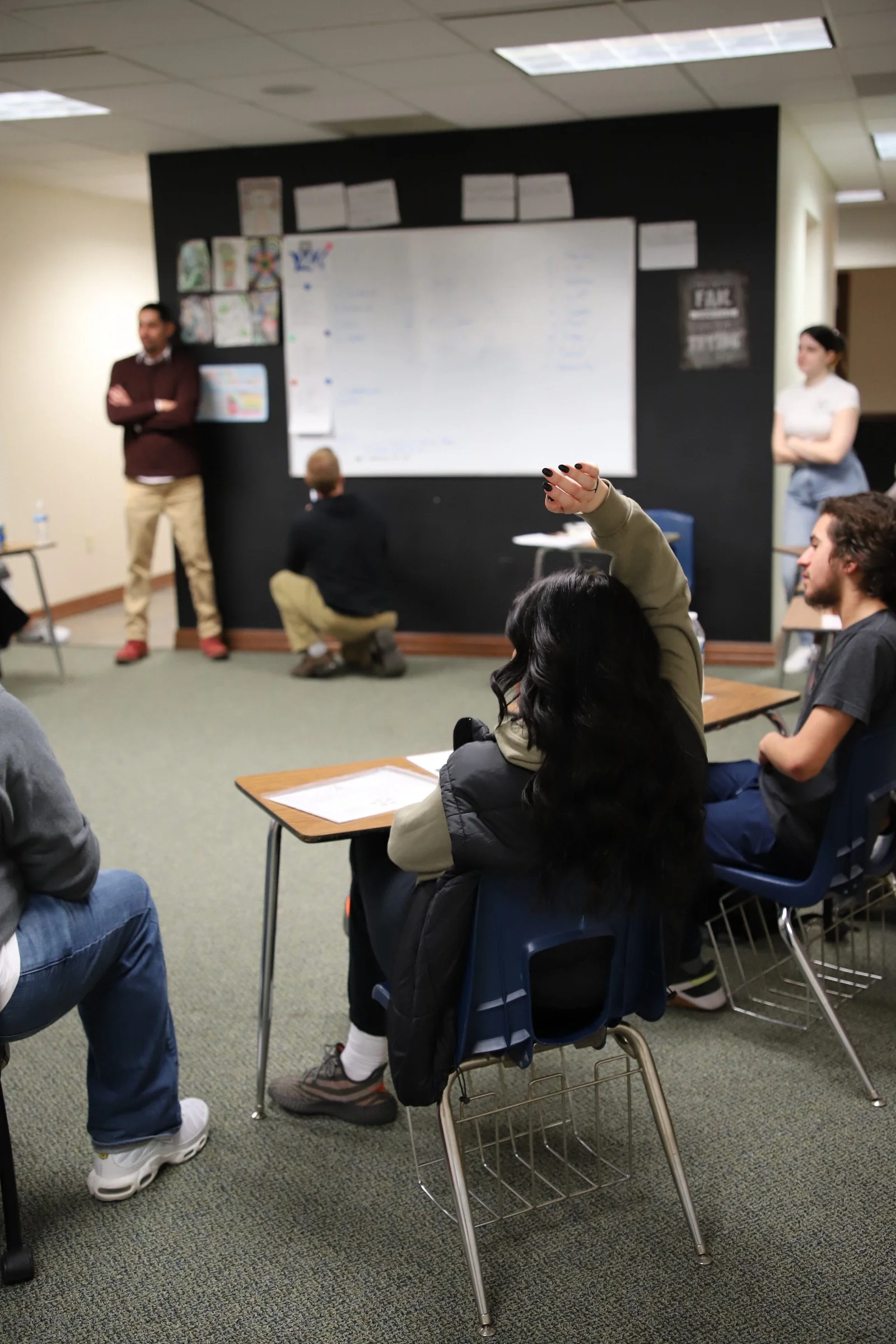 A classroom or meeting room with students listening to a presentation by two instructors at the front near a whiteboard.