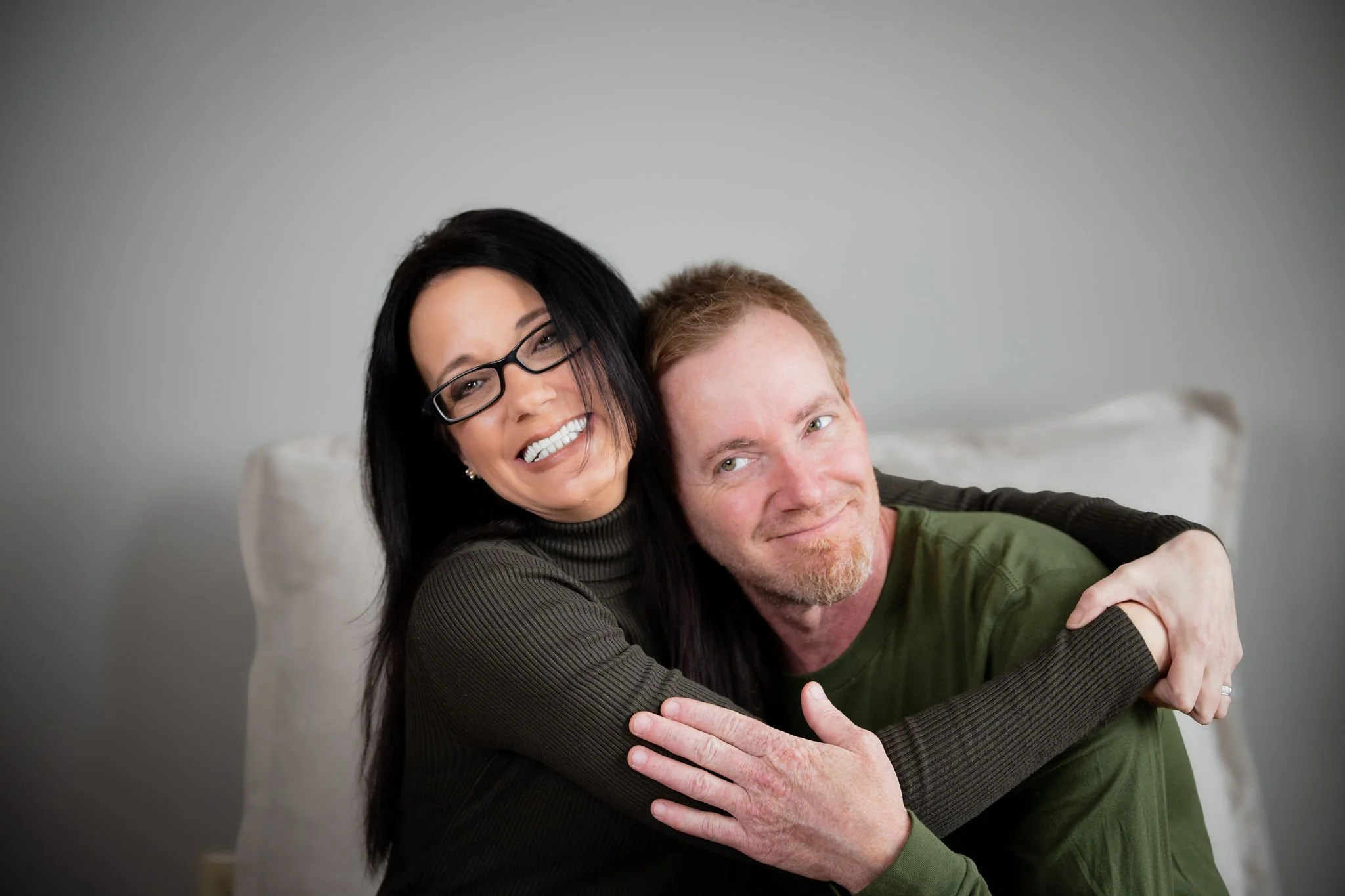 A woman with black hair, glasses, and a brown turtleneck hugging a man with red hair and a green shirt, both smiling and sitting on a sofa.