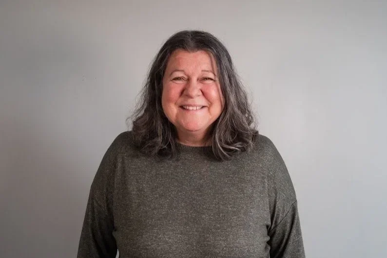 Smiling woman with shoulder-length gray hair wearing a dark gray sweater standing against a plain light gray wall.