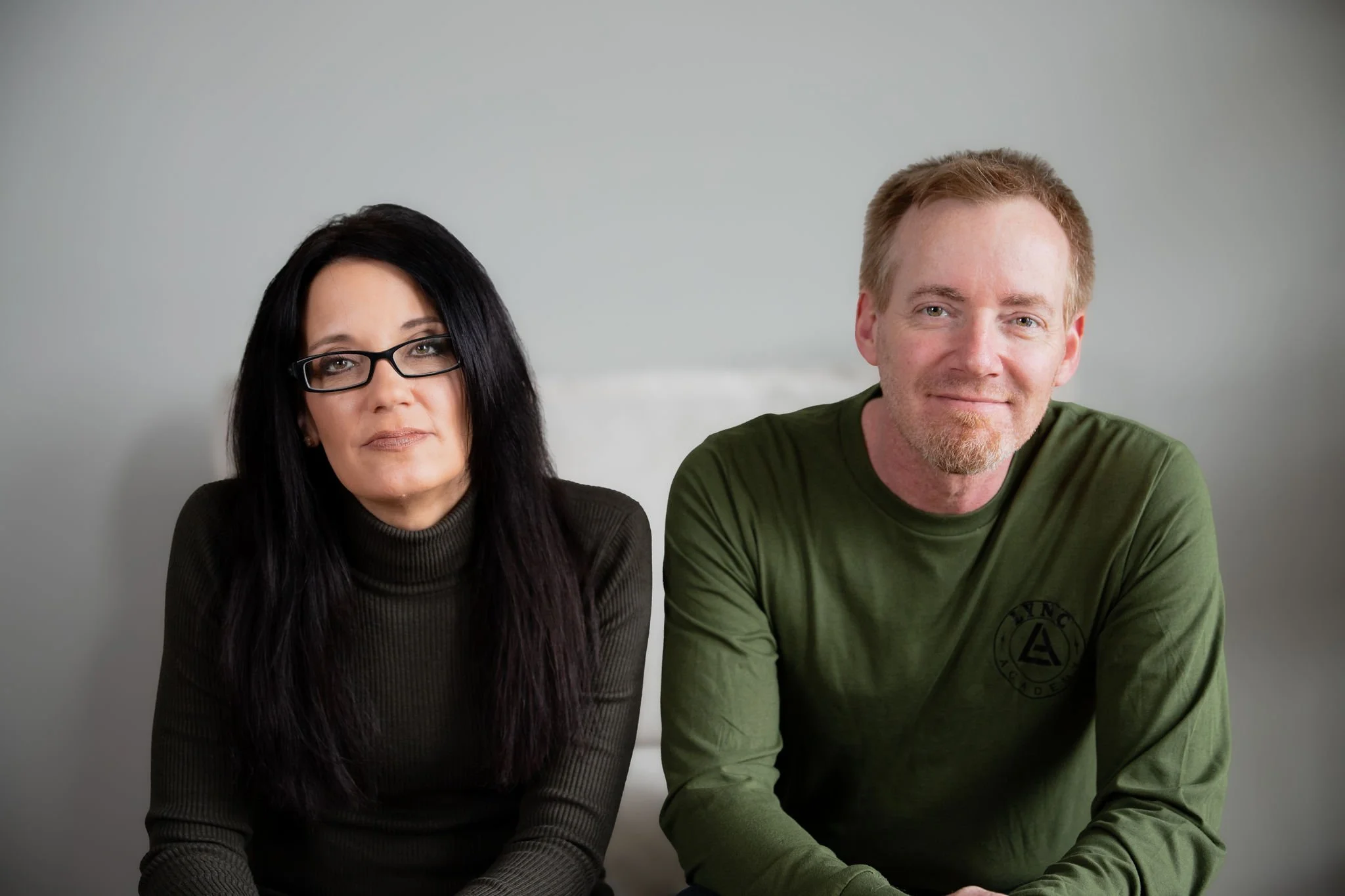 A woman with long dark hair, glasses, and a dark green turtleneck sitting next to a smiling man with short light hair, a beard, and a green long-sleeve shirt.