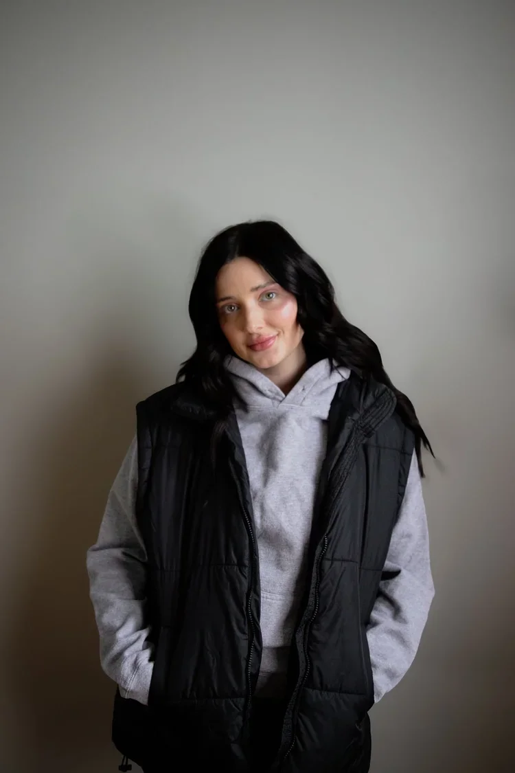 Young woman with black hair, wearing a gray hoodie and black vest, standing against a plain light gray wall.