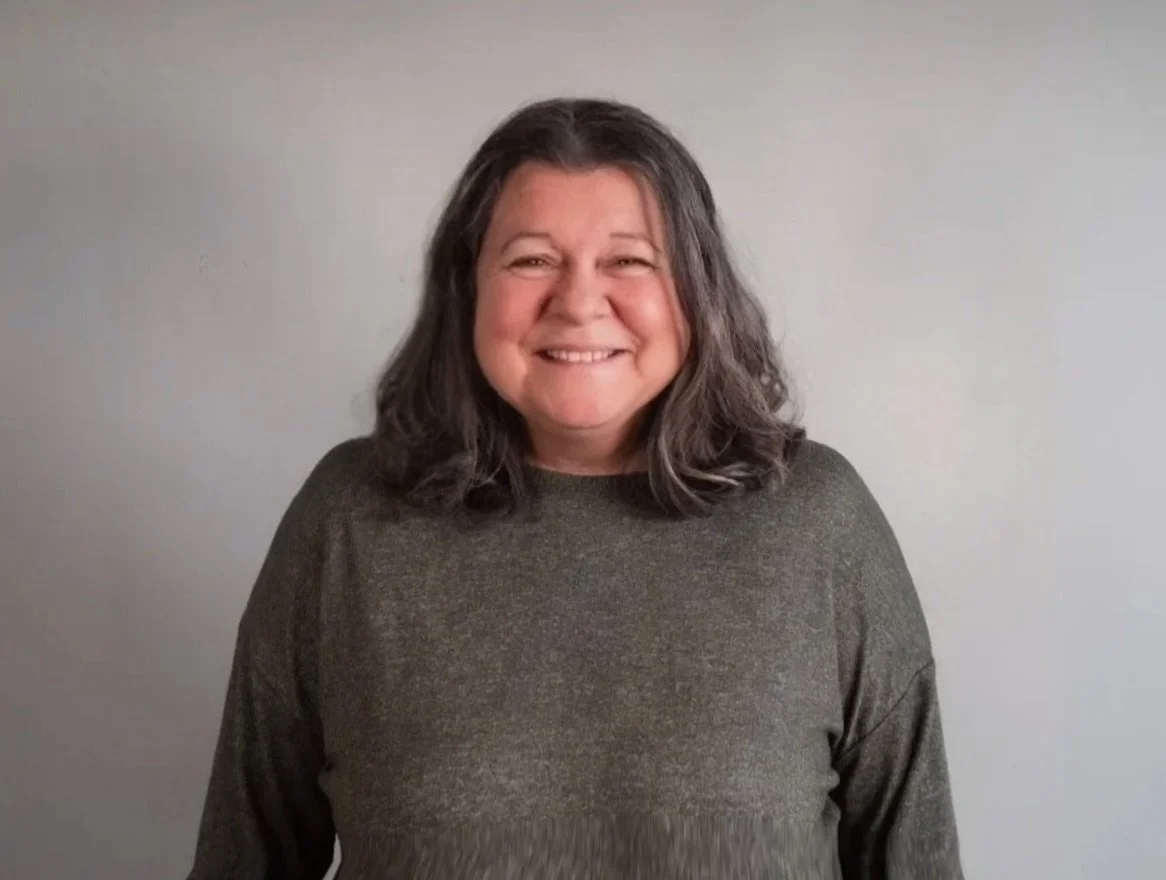 Smiling woman with shoulder-length gray hair wearing a dark gray sweater standing against a plain light gray wall.