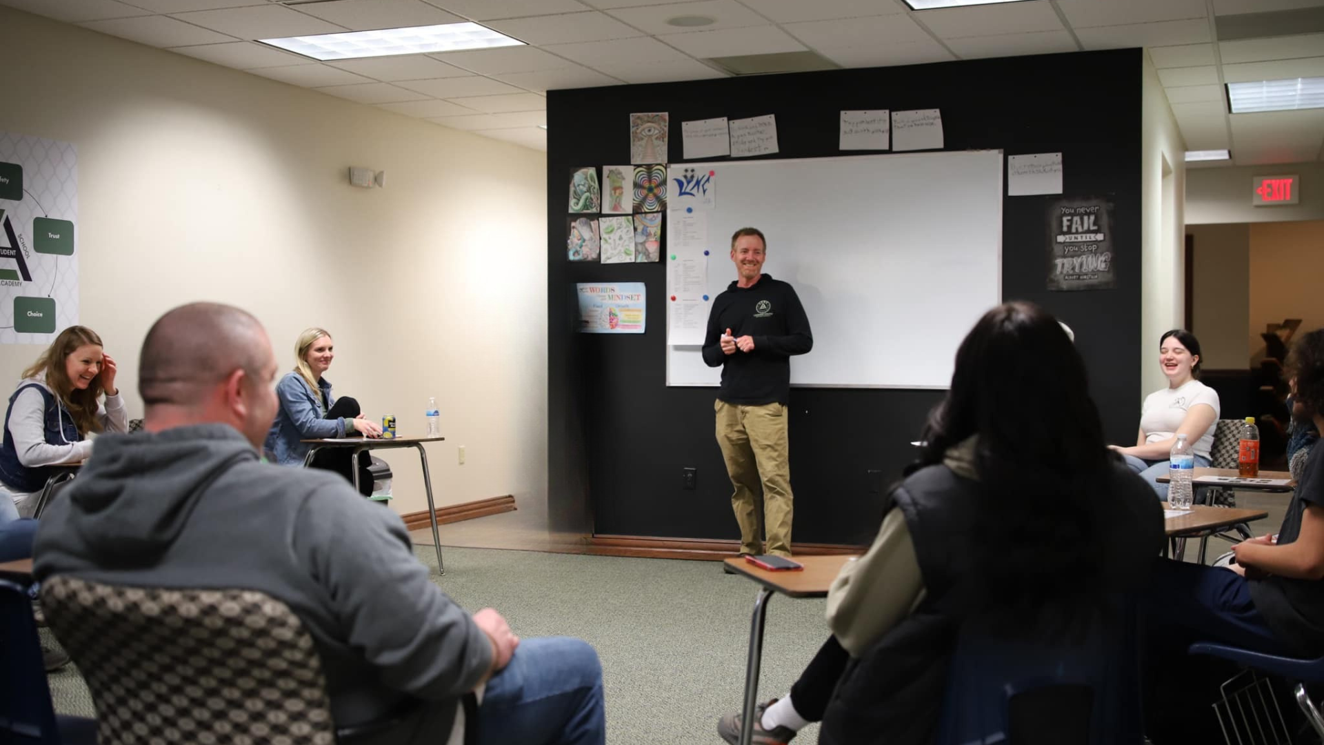 A classroom with students sitting at desks and a teacher standing at the front near a whiteboard, smiling and addressing the class.