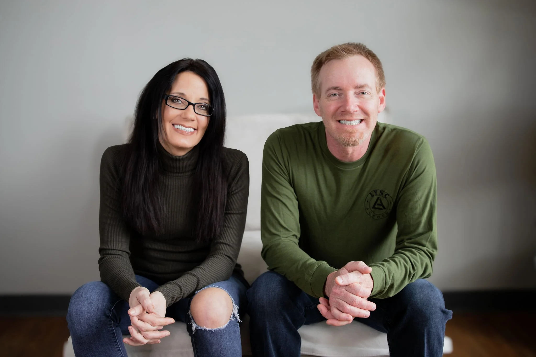 A woman with long black hair, glasses, a dark turtleneck, and ripped jeans, sitting next to a man with short red hair, a beard, a green long sleeve shirt, and dark jeans, both smiling and sitting on a white couch in front of a plain wall.
