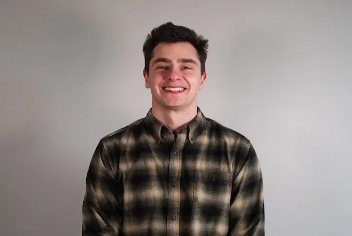 Young man smiling, wearing a plaid shirt, standing against a plain white wall.