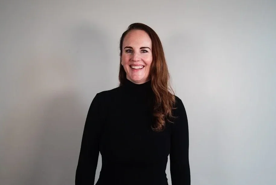 A woman with brown hair wearing a black turtleneck, smiling, standing against a plain light-colored wall.