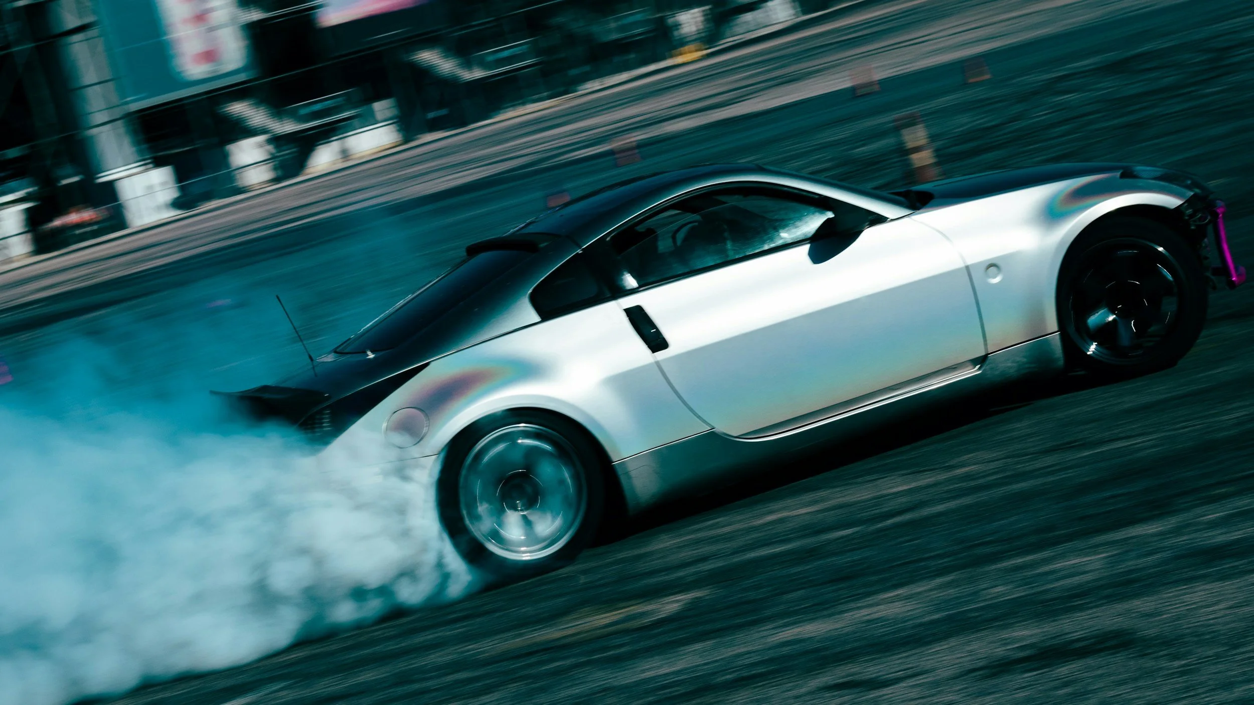 A silver sports car drifting on a city street, smoke coming from the tires, with blurred buildings in the background.