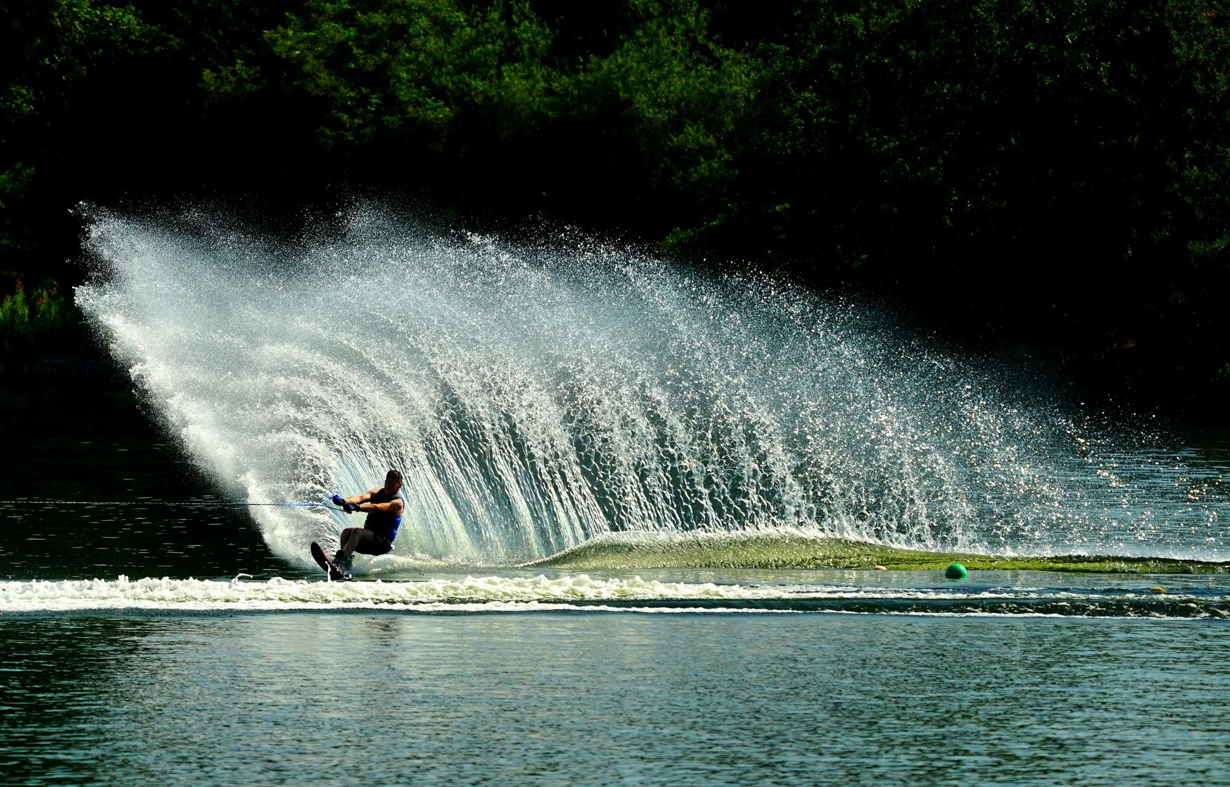 A person water skiing on a lake, creating a large splash and wake, with a wooded shoreline in the background.