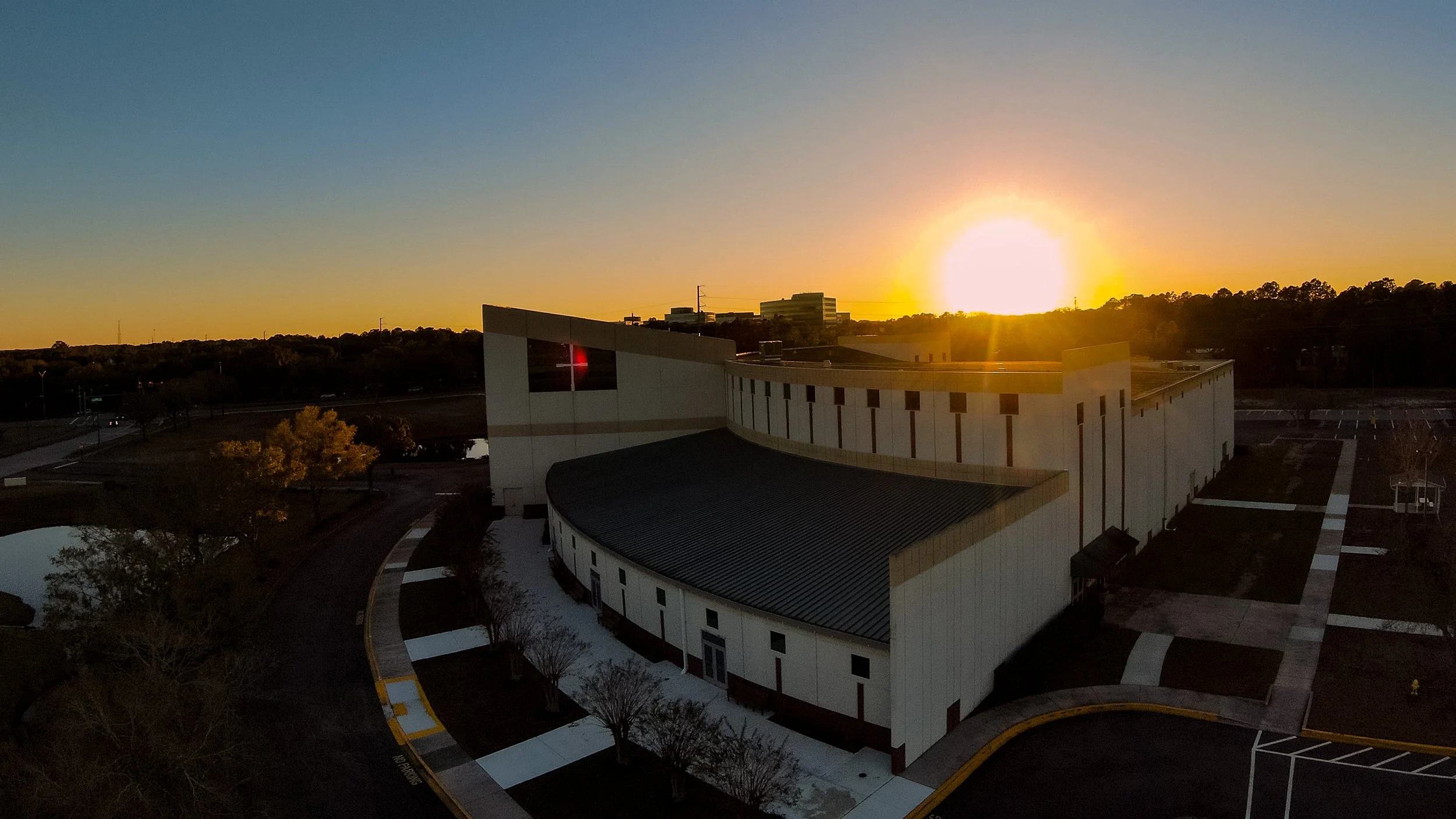 A modern building at sunset with a clear sky, trees, and a parking lot in the foreground.