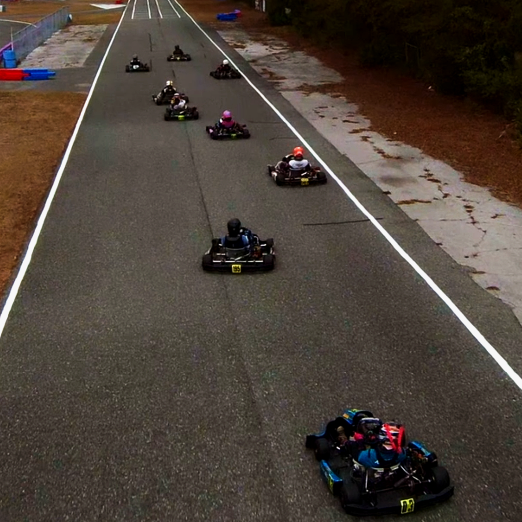 Multiple go-karts racing on a paved track, with drivers wearing helmets and racing suits.
