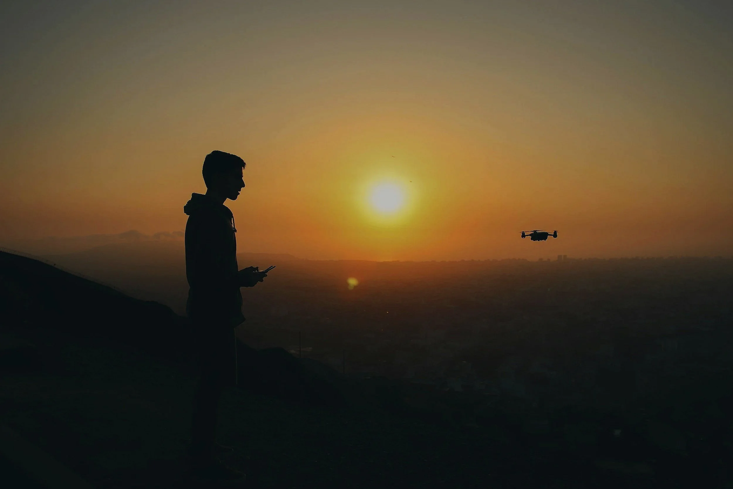 Silhouette of a person holding a phone, standing on a hill during sunset, with a drone flying in the sky.