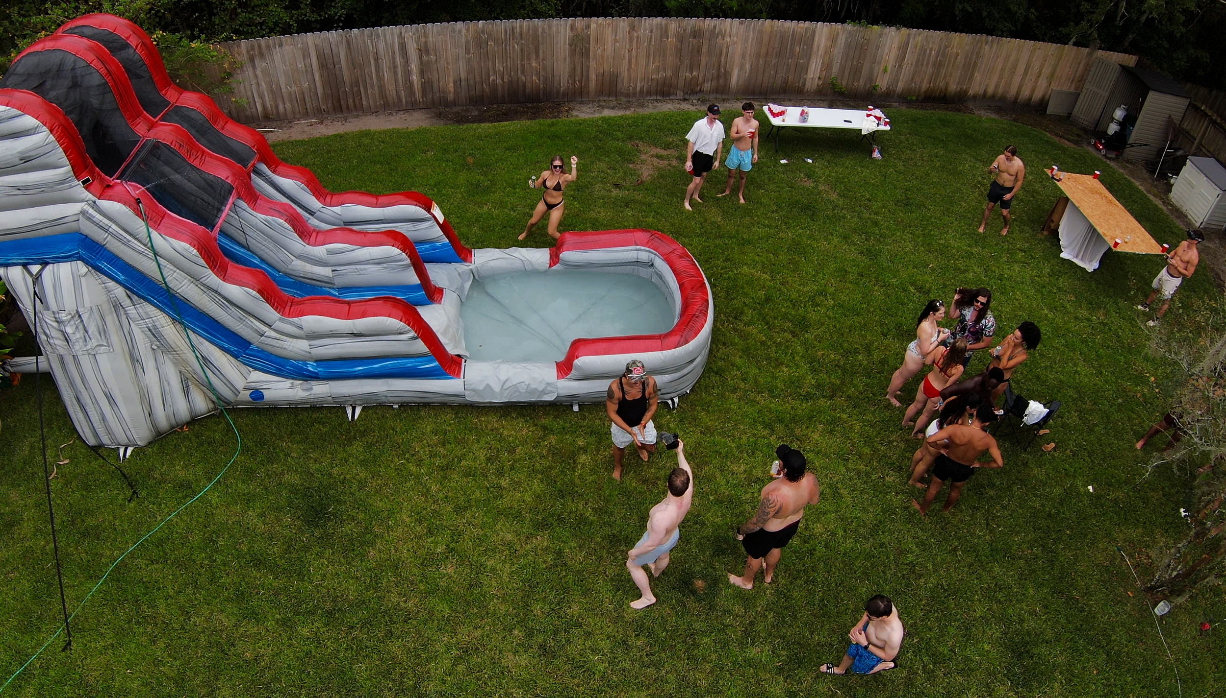 A backyard party with a large inflatable water slide, people in swimsuits, and some standing and chatting on the grass. There are tables and a fence in the background.