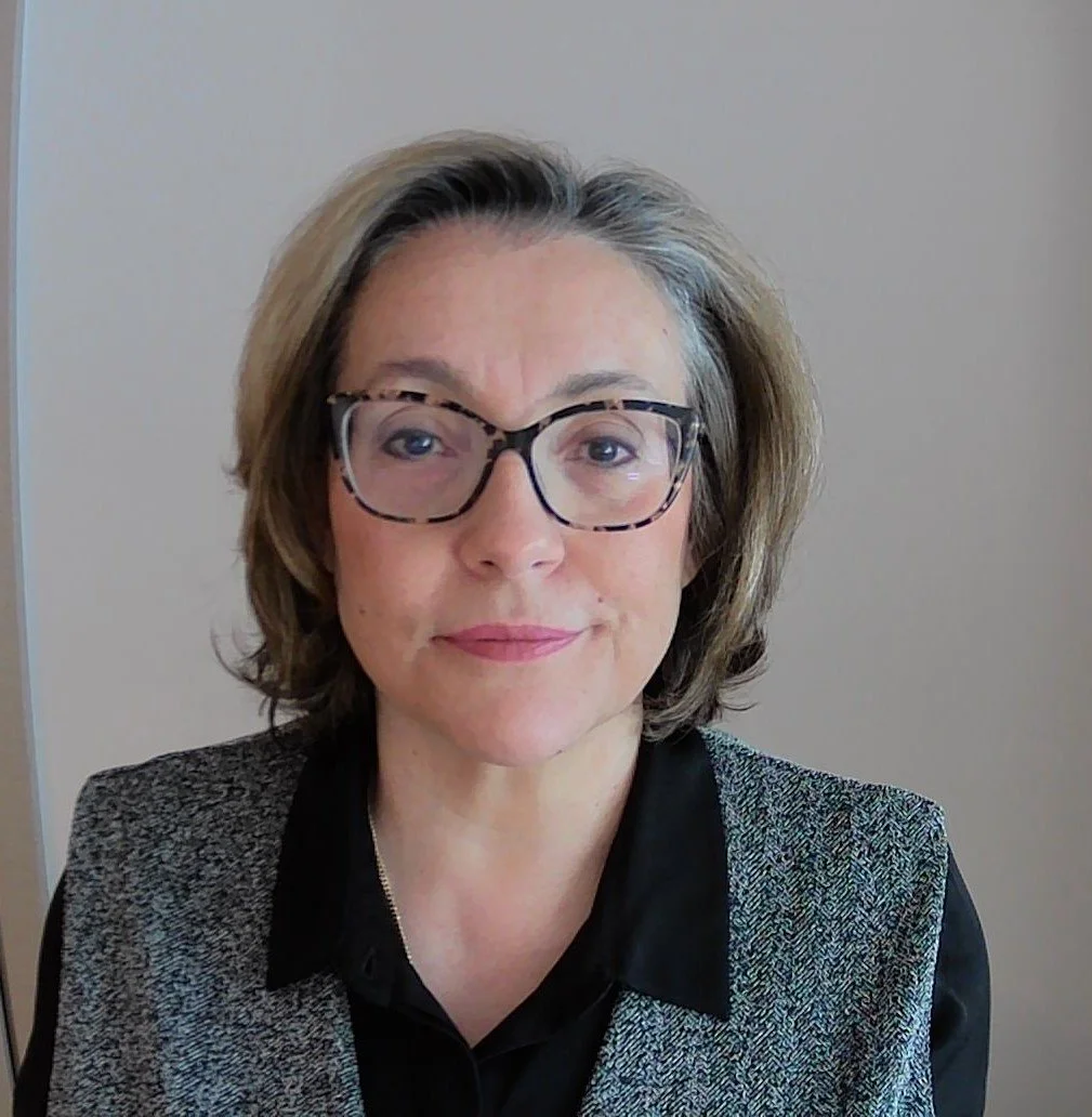 A woman with shoulder-length gray hair wearing glasses and a black shirt with a gray blazer, sitting in front of a plain beige wall.