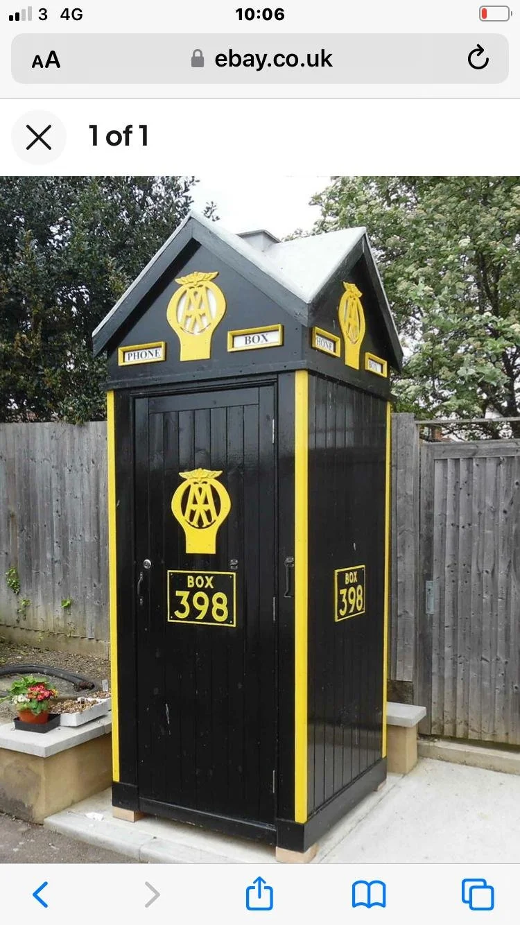 A black and yellow postal box labeled 'Box 398' with the Masonic symbol on it, located outdoors next to a wooden fence and small flower bed.