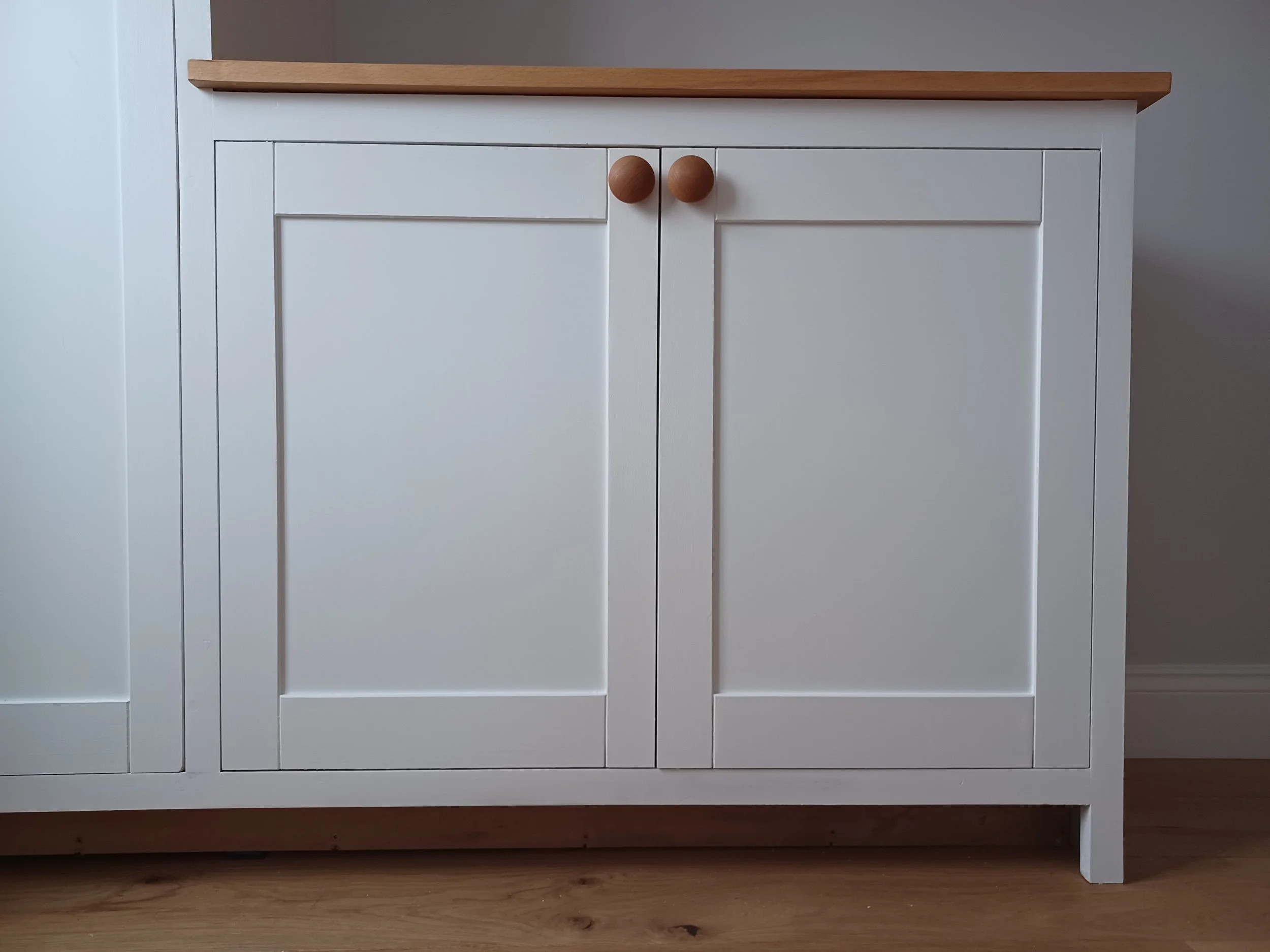 A white wooden cabinet with two doors, wooden knobs, and a wooden top surface, set against a light-colored wall and placed on a wooden floor.