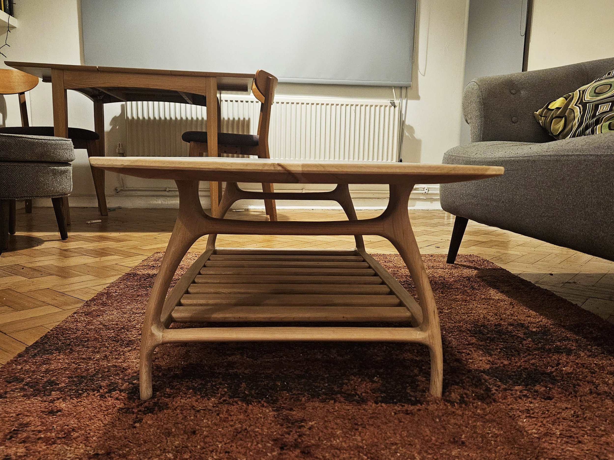 A low wooden coffee table with slatted shelf in a living room, with a reddish-brown rug underneath, surrounded by a gray sofa with patterned pillows, a wooden dining table with chairs, and a window with a blue shade.