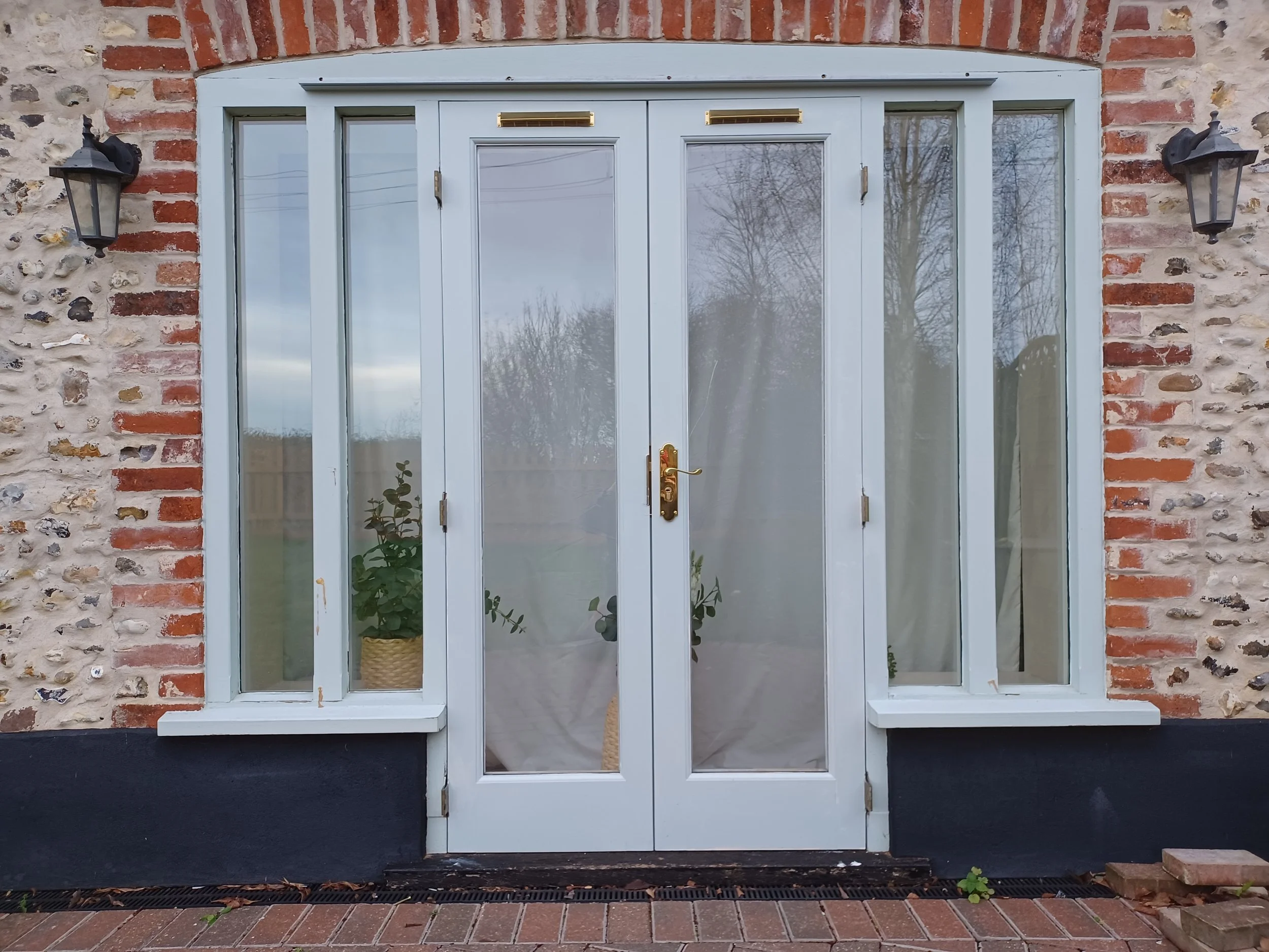 White glass-paneled door and windows set in a brick and stone exterior wall, with outdoor lamps on either side and potted plants visible through the glass.