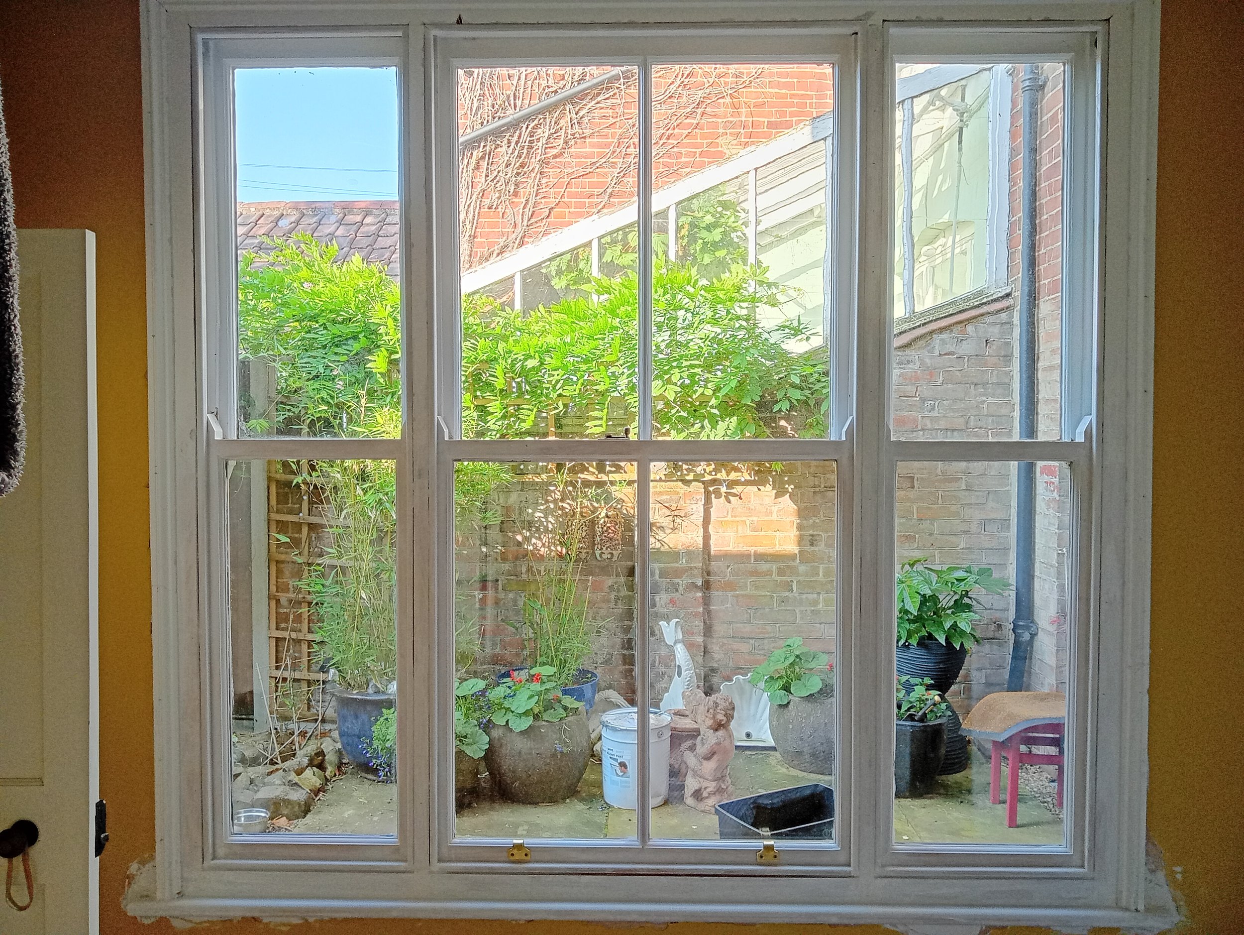 View of a small backyard garden through a large window with white framing. The garden has potted plants and garden ornaments, with a brick wall and stairs in the background.