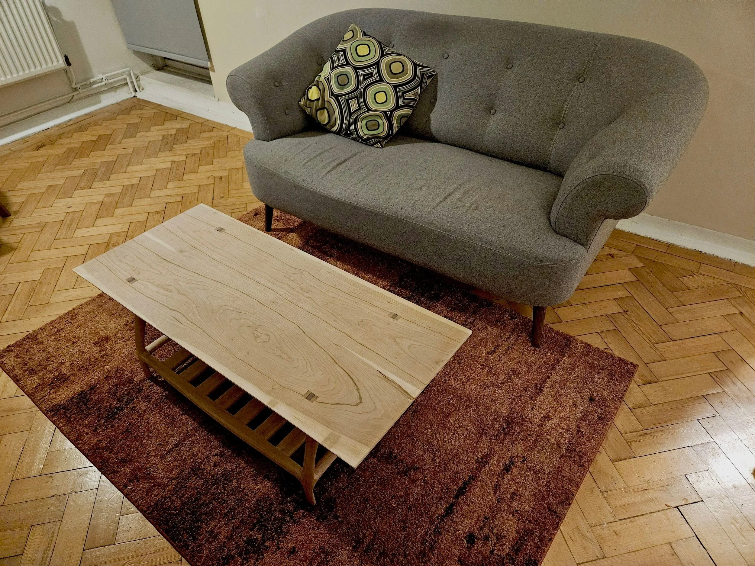 A gray upholstered sofa with a patterned throw pillow, a light wood coffee table with a slatted shelf underneath, on a red-brown rug, in a room with wooden parquet flooring.