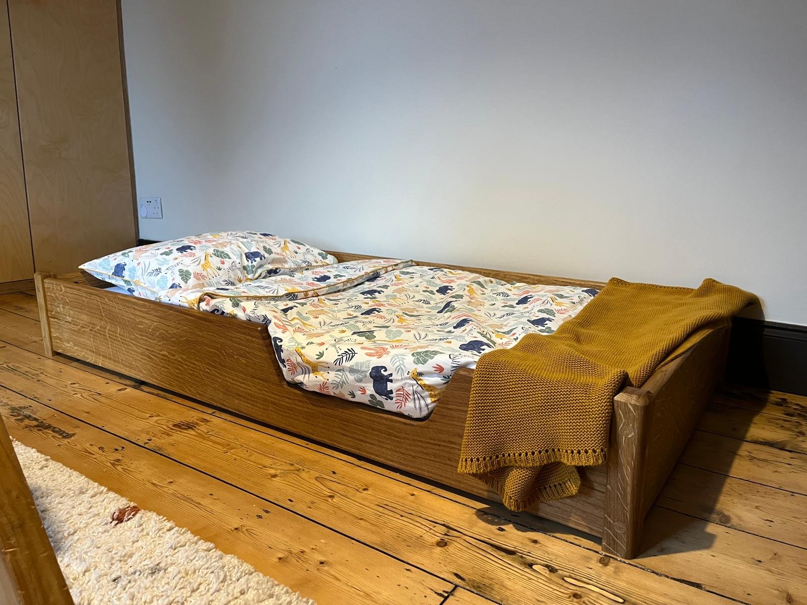 A wooden bed with colorful patterned bedding and a mustard yellow blanket in a minimally decorated room with wooden floor and light-colored walls.