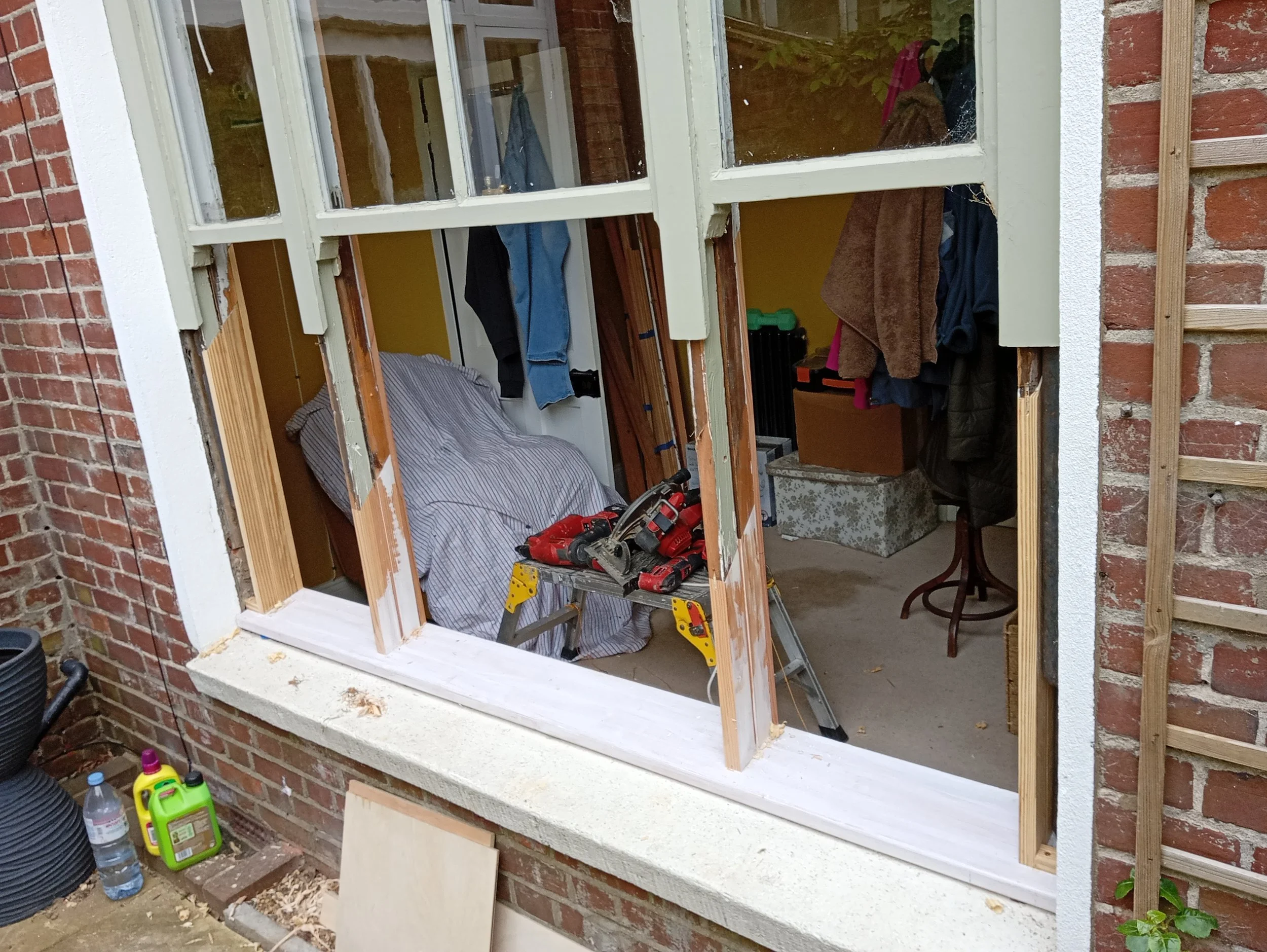 A window is being renovated, with the glass panes removed, revealing tools and construction materials inside. Outside, on the ground, there are bottles of water and cleaning supplies.