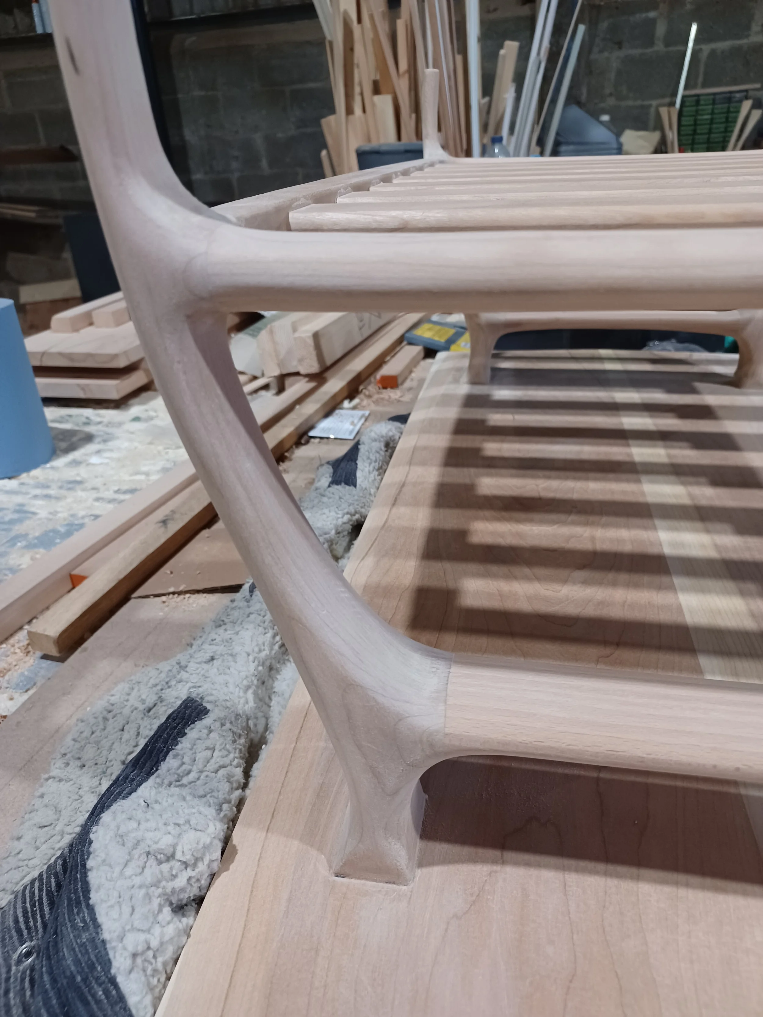 Close-up of an unfinished wooden chair with smooth, curved legs and horizontal slats for the seat, on a woodworking workbench with other wood pieces and tools in the background.