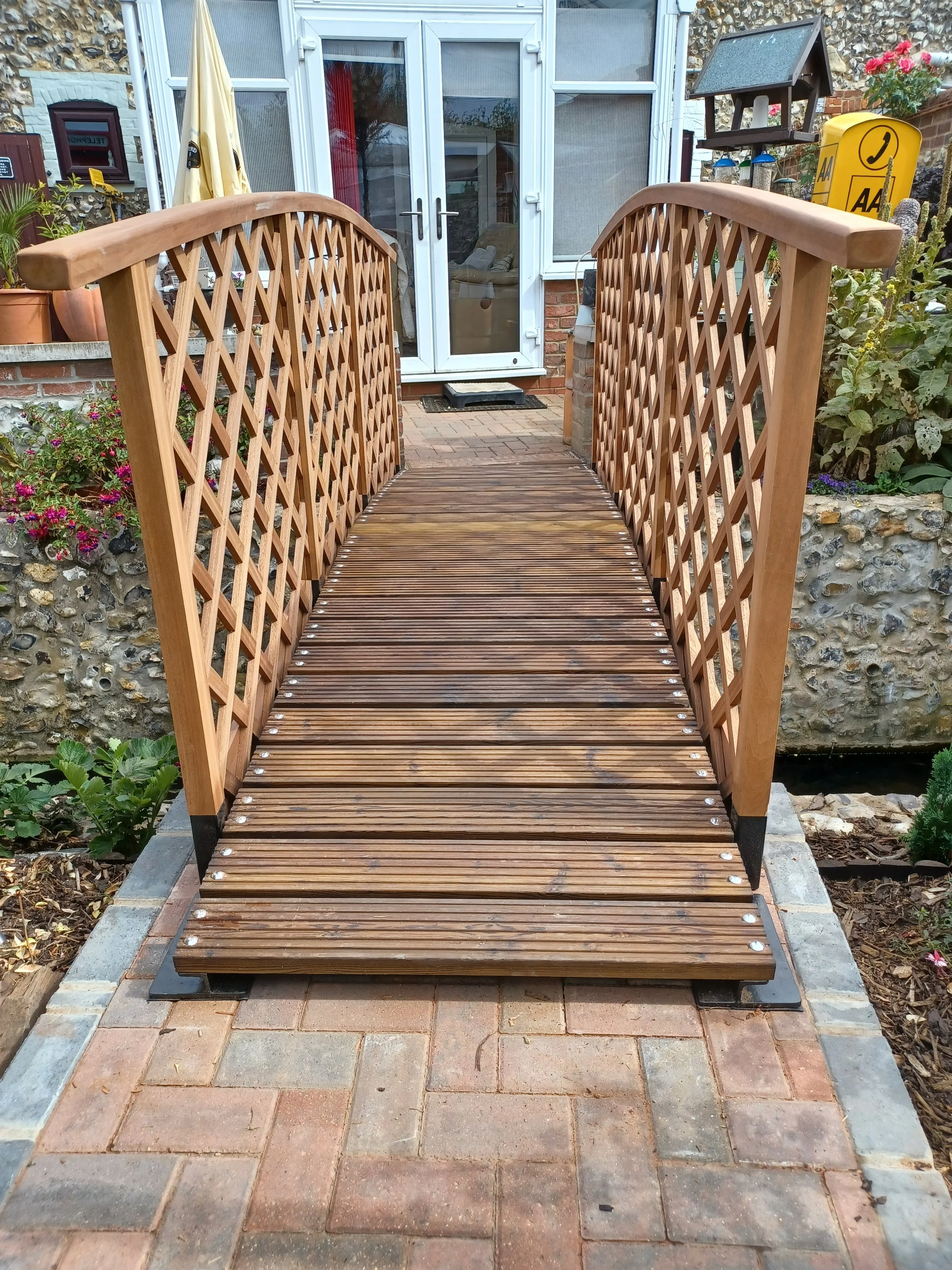 Wooden garden bridge with lattice sides spanning over a small garden path entrance to a house door.