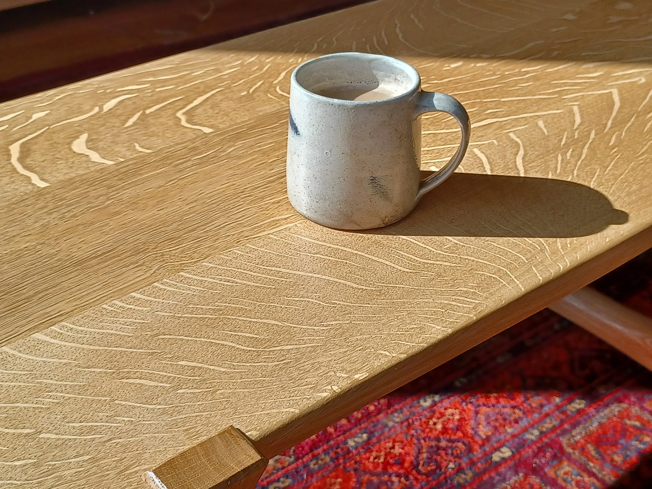 A ceramic mug filled with a beverage, placed on a wooden tabletop with textured grain and carved patterns, with a shadow cast to the right.