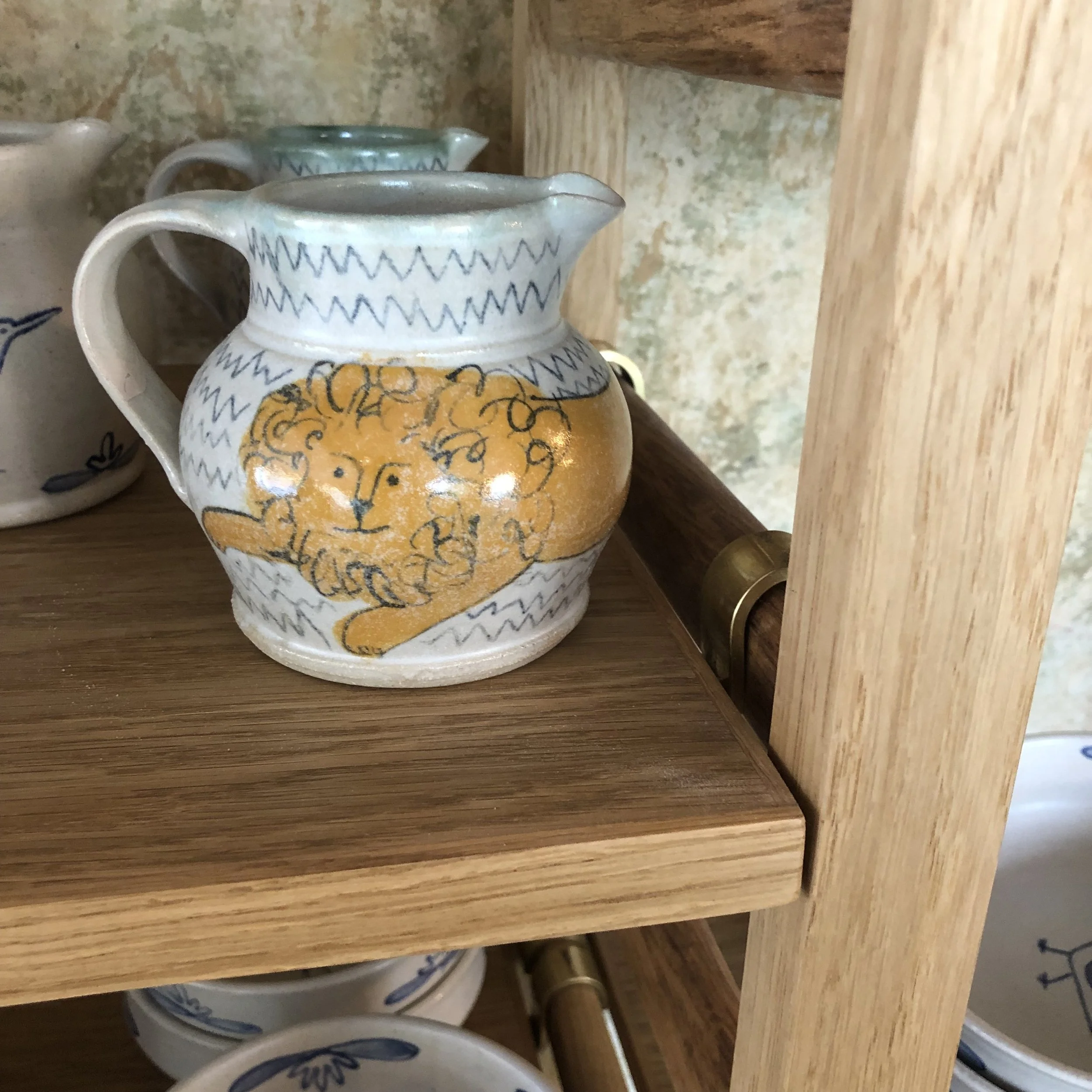 A ceramic pitcher with a lion illustration on it, sitting on a wooden shelf.