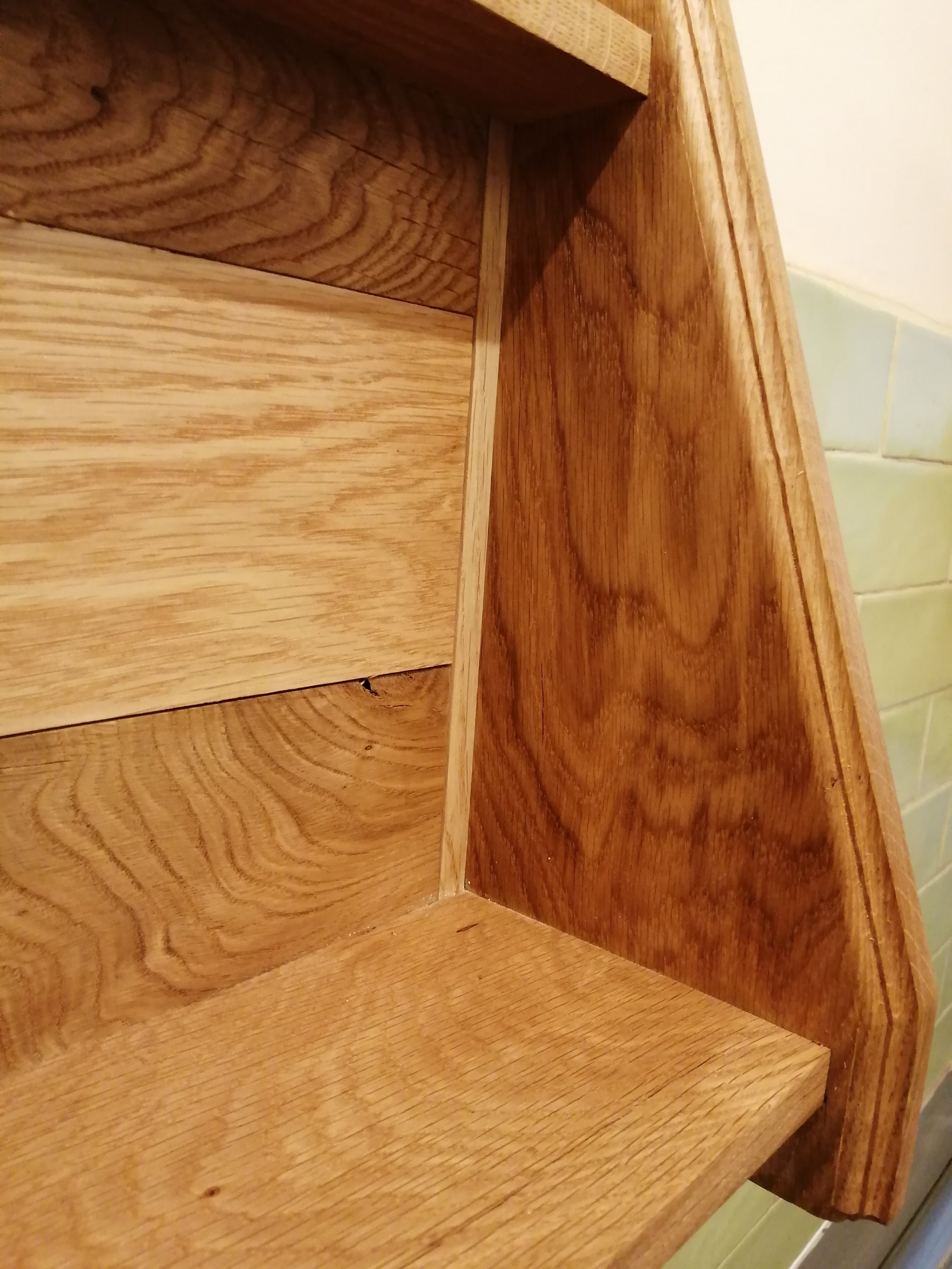 Close-up view of a wooden shelf showing the grain and joints of the wood with a tiled wall in the background.