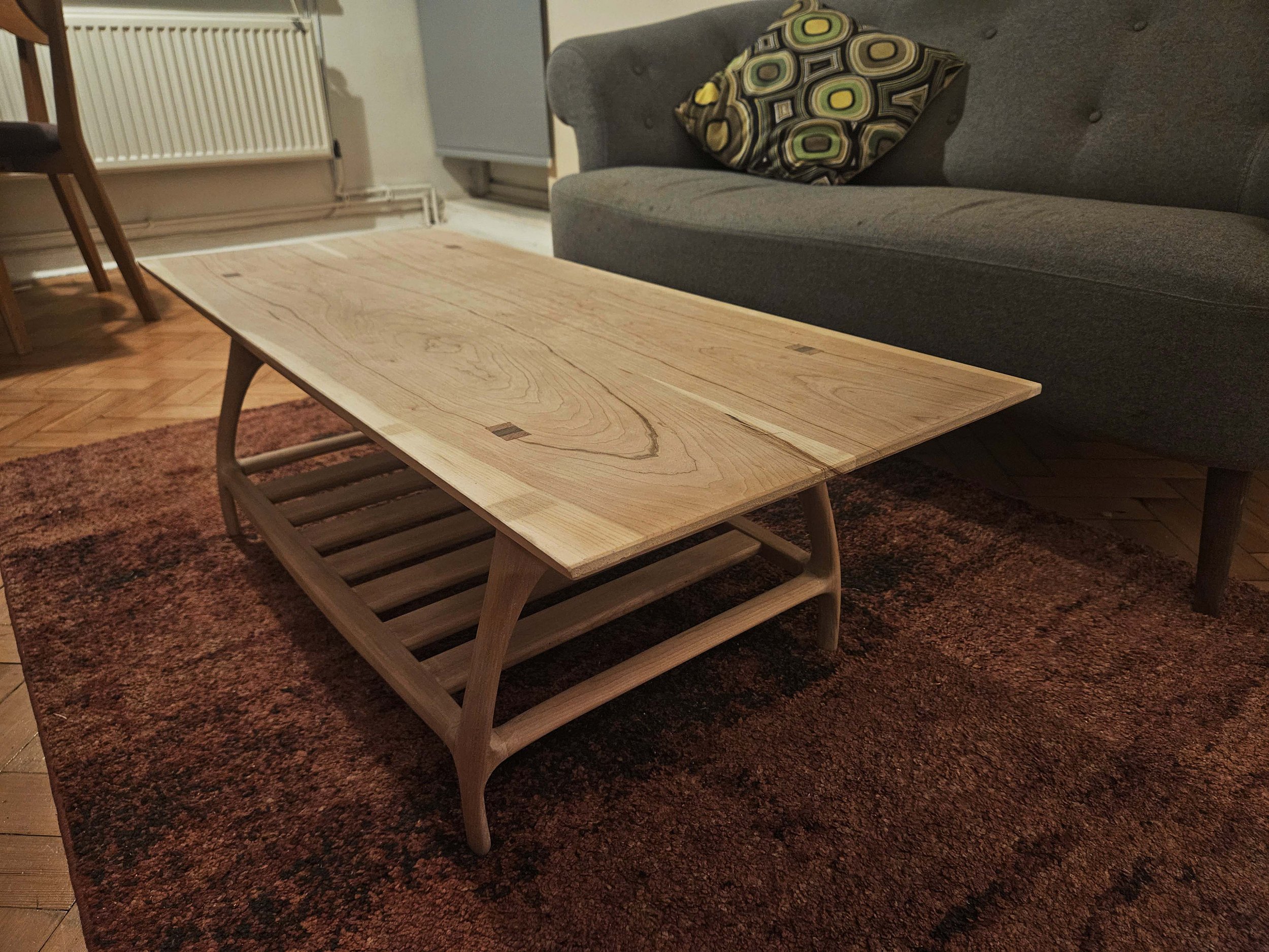 Wooden coffee table with a slatted lower shelf on a reddish-brown rug in front of a gray sofa with a patterned pillow.