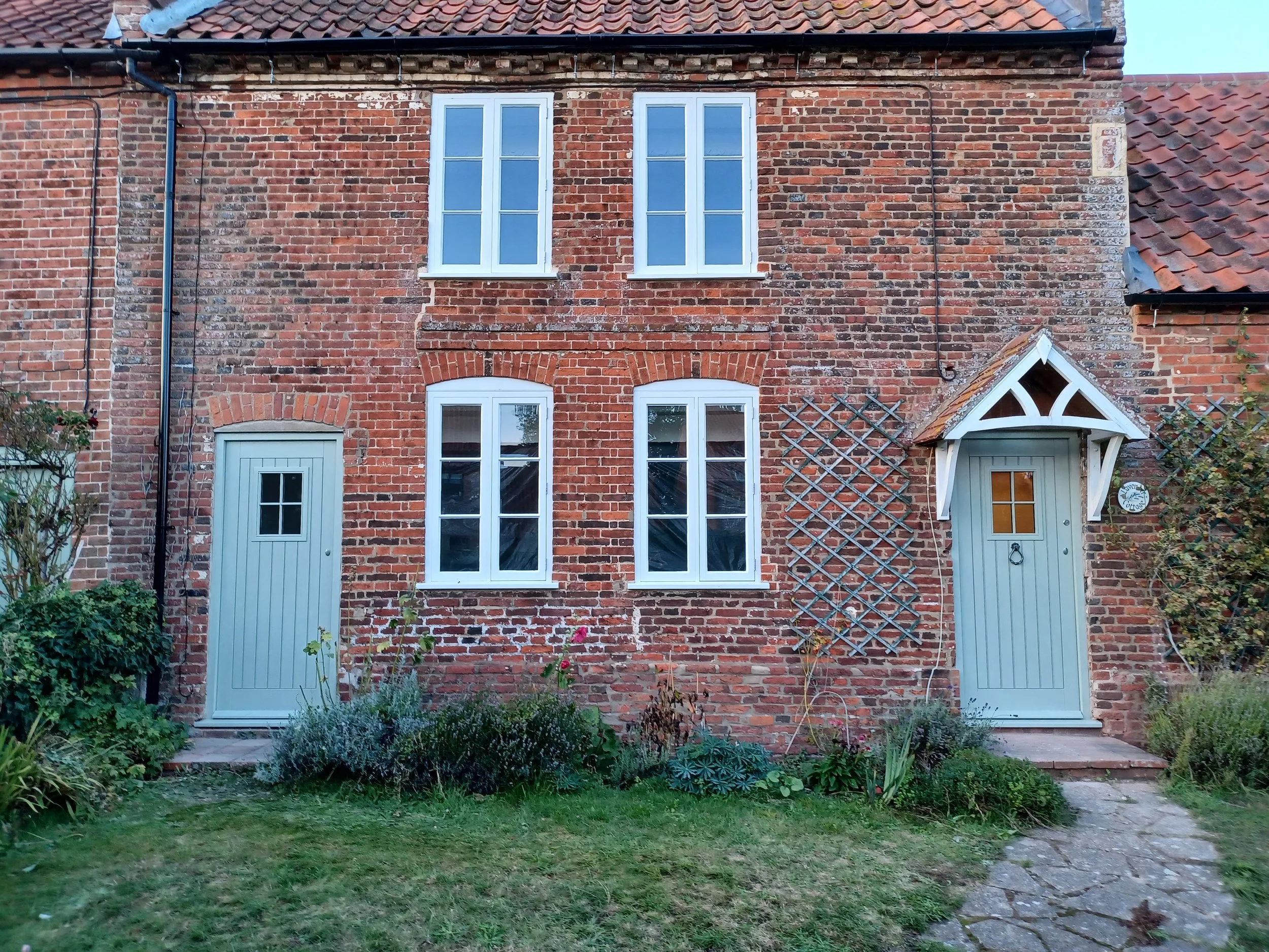 Brick house with two stories, four white-framed windows, two blue doors, and a small garden with plants in front.