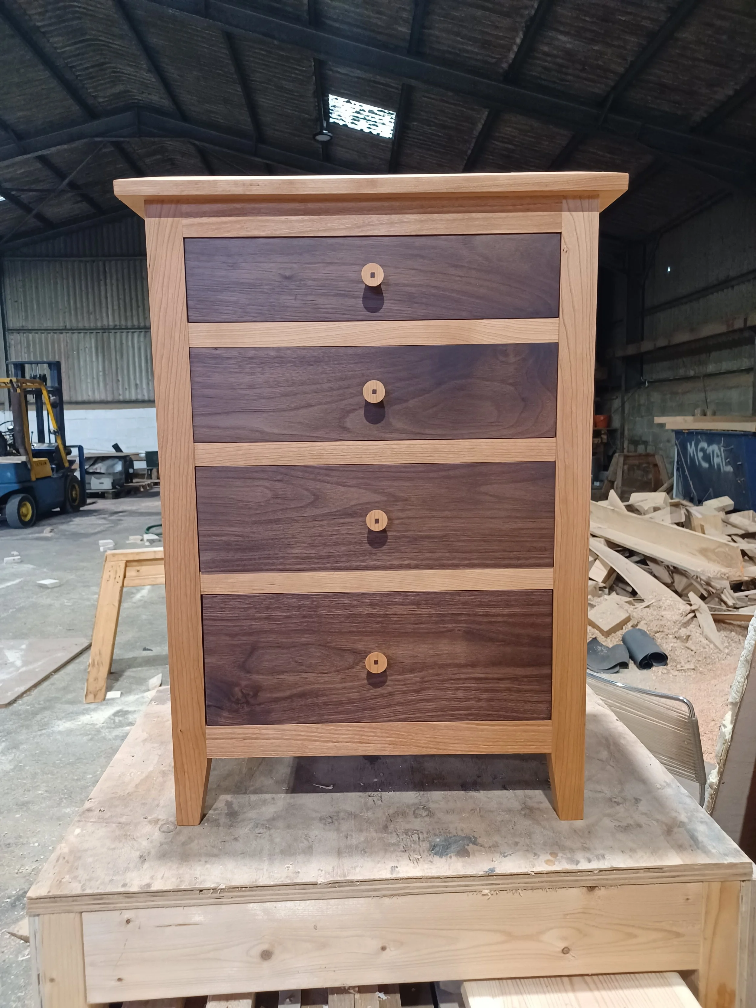 A wooden chest of drawers with four dark wood drawers and round wooden knobs, situated on a workbench in a woodworking workshop.