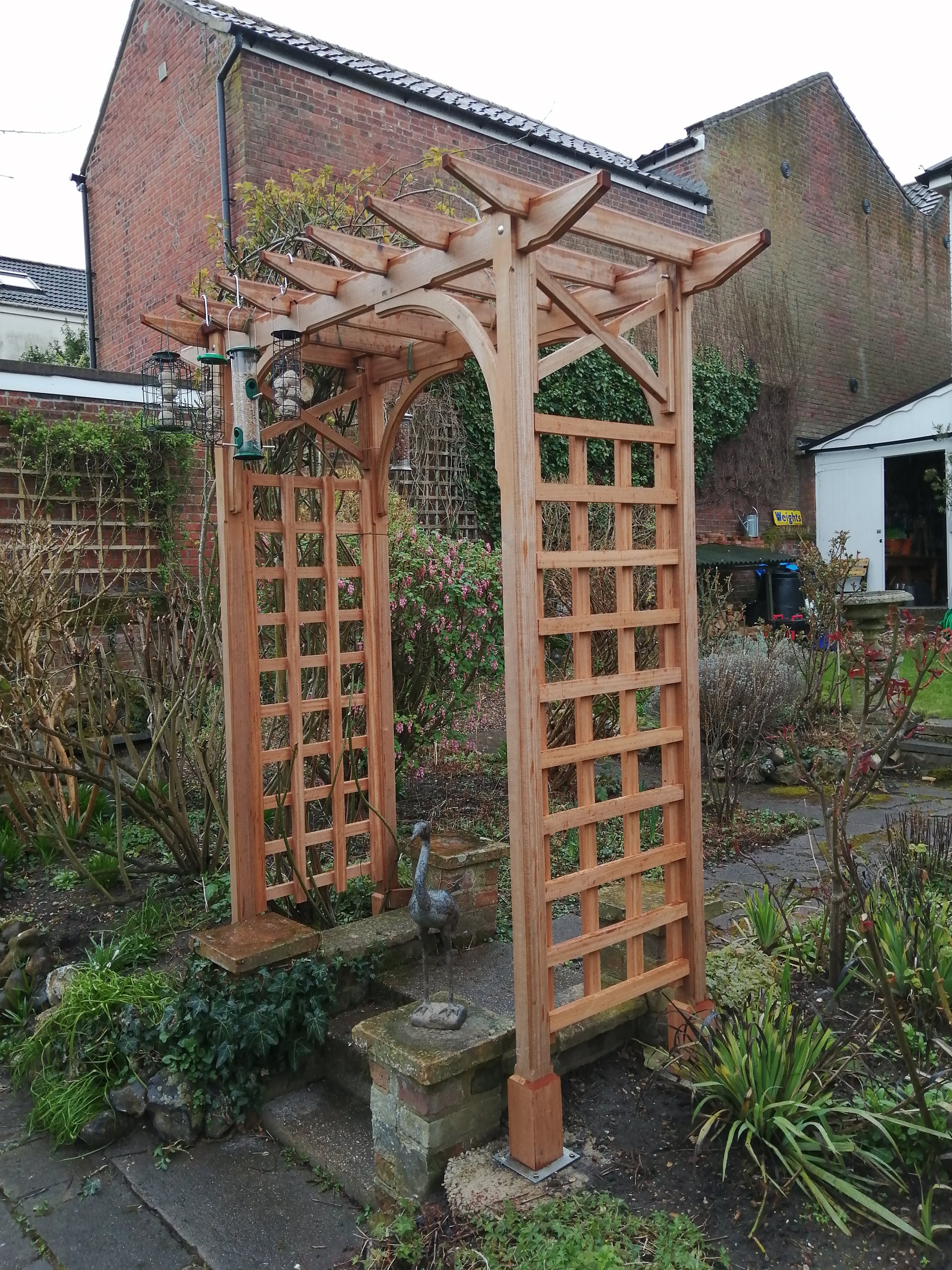 Wooden garden arch with lattice sides and roof, with decorations and plants around, in a backyard garden.