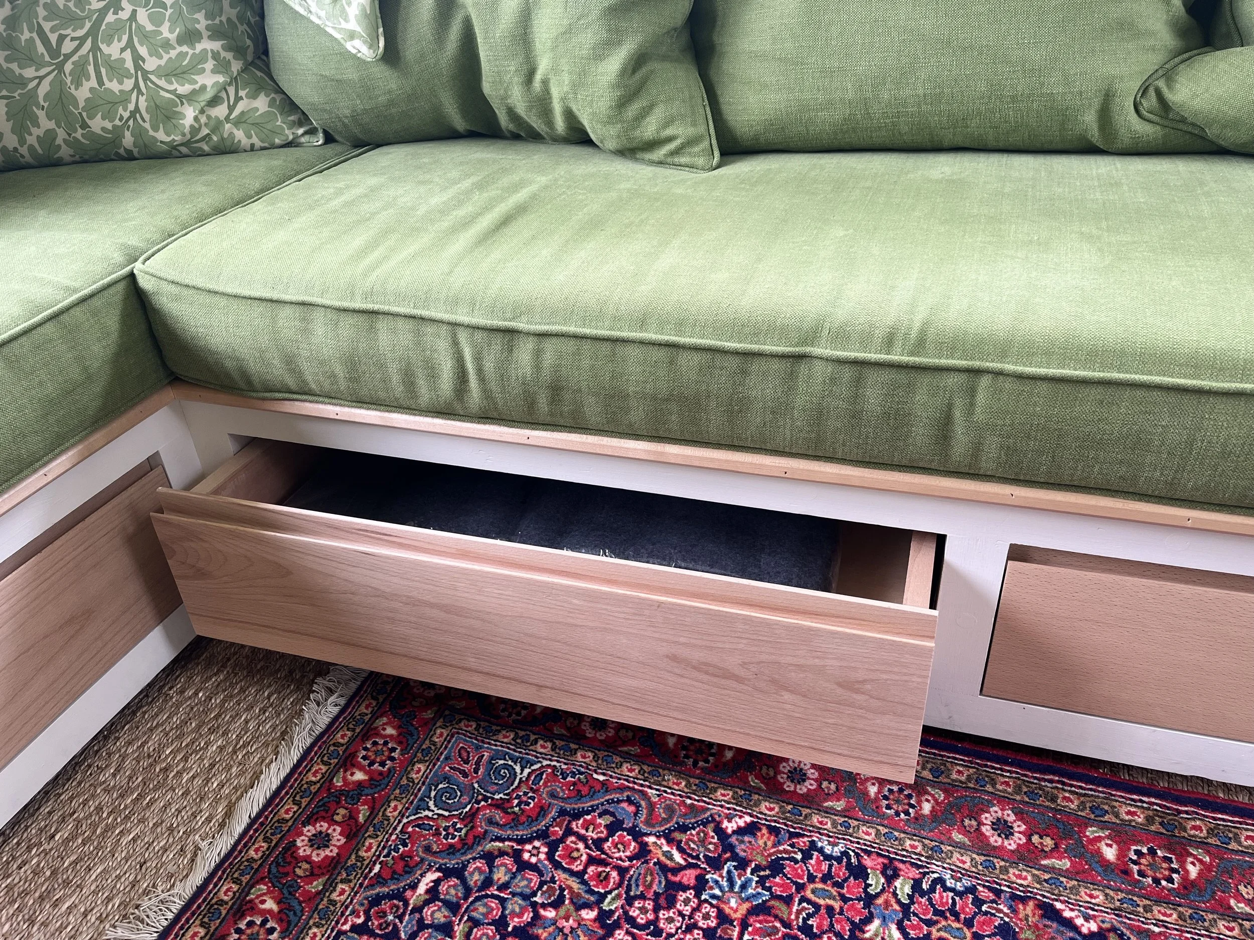 Open drawer underneath a green sofa with patterned pillows, revealing a black item inside a room with a patterned area rug.