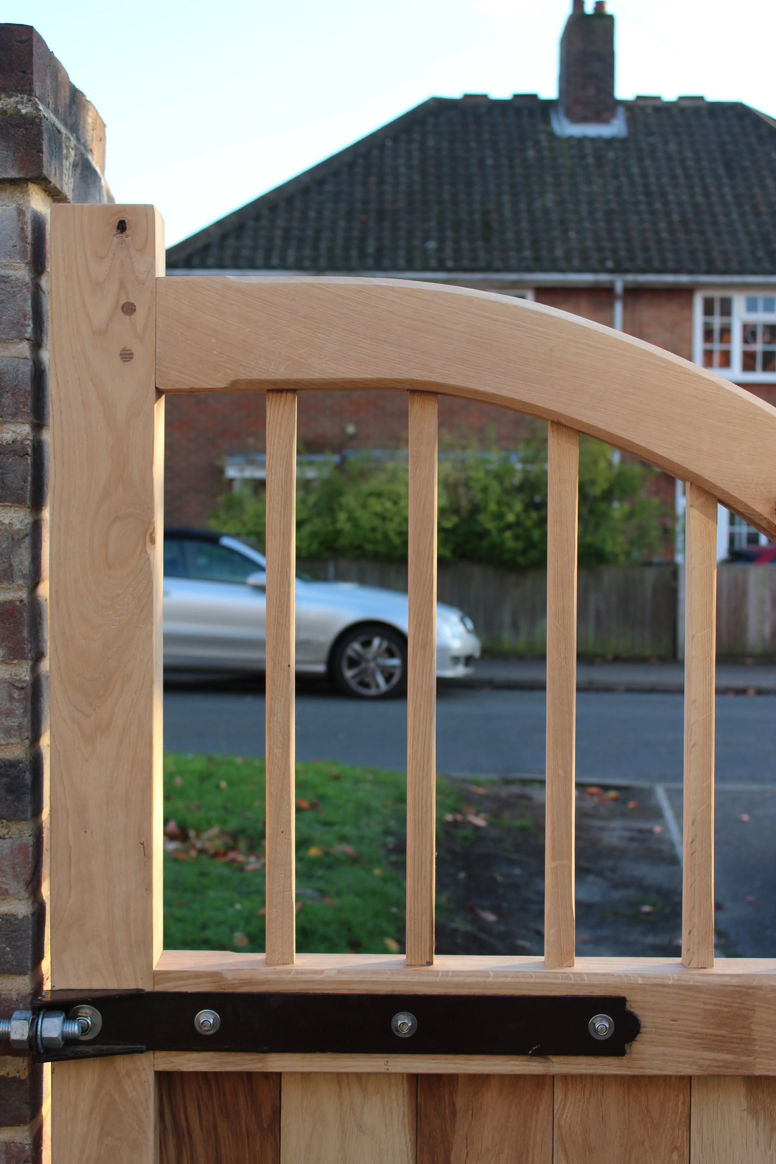 A close-up of a wooden gate with vertical slats, mounted on a brick wall with metal hardware, with a residential street and house in the background.