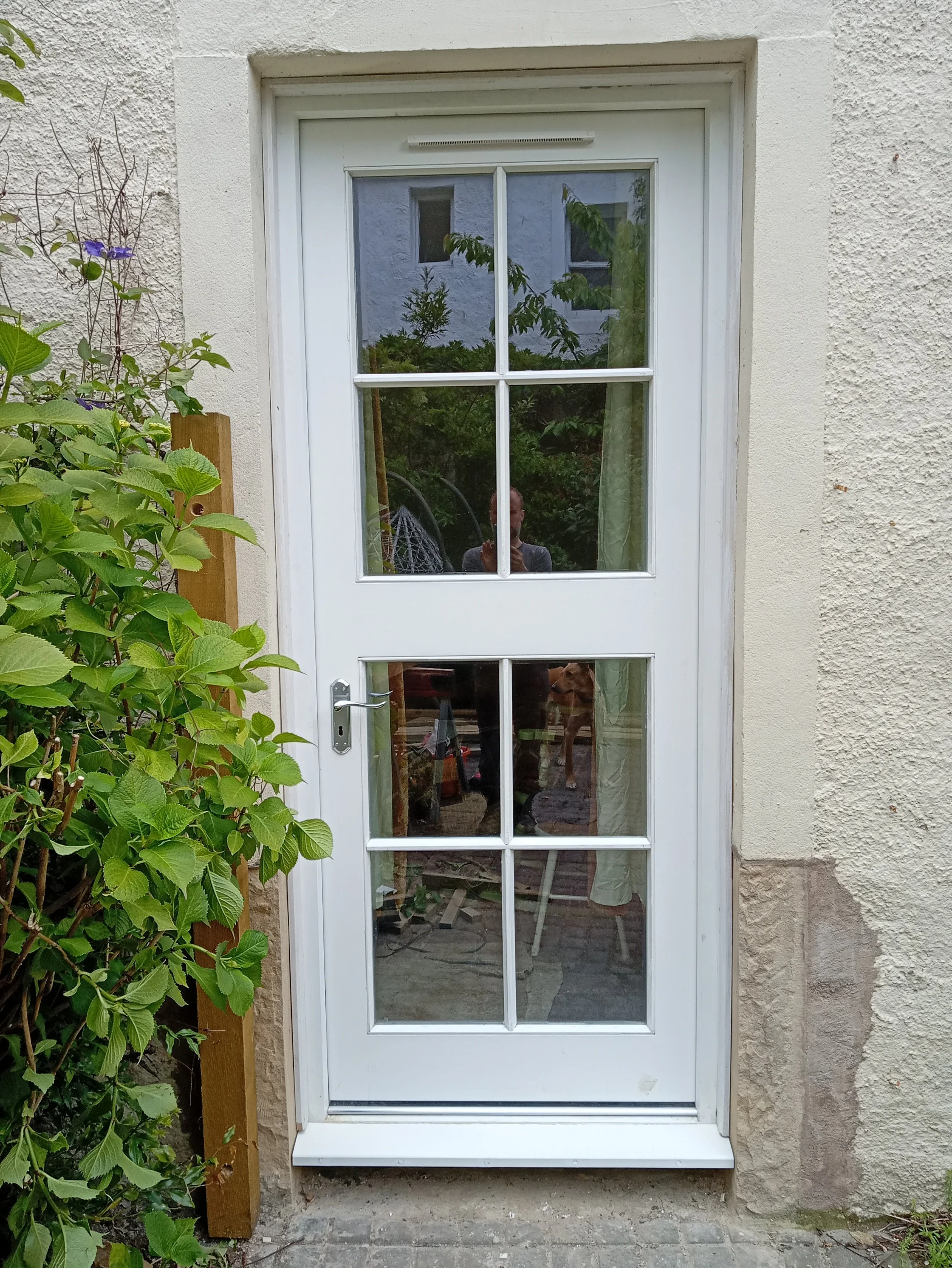White exterior door with multiple glass panes, reflecting person, greenery, and garden furniture outside.