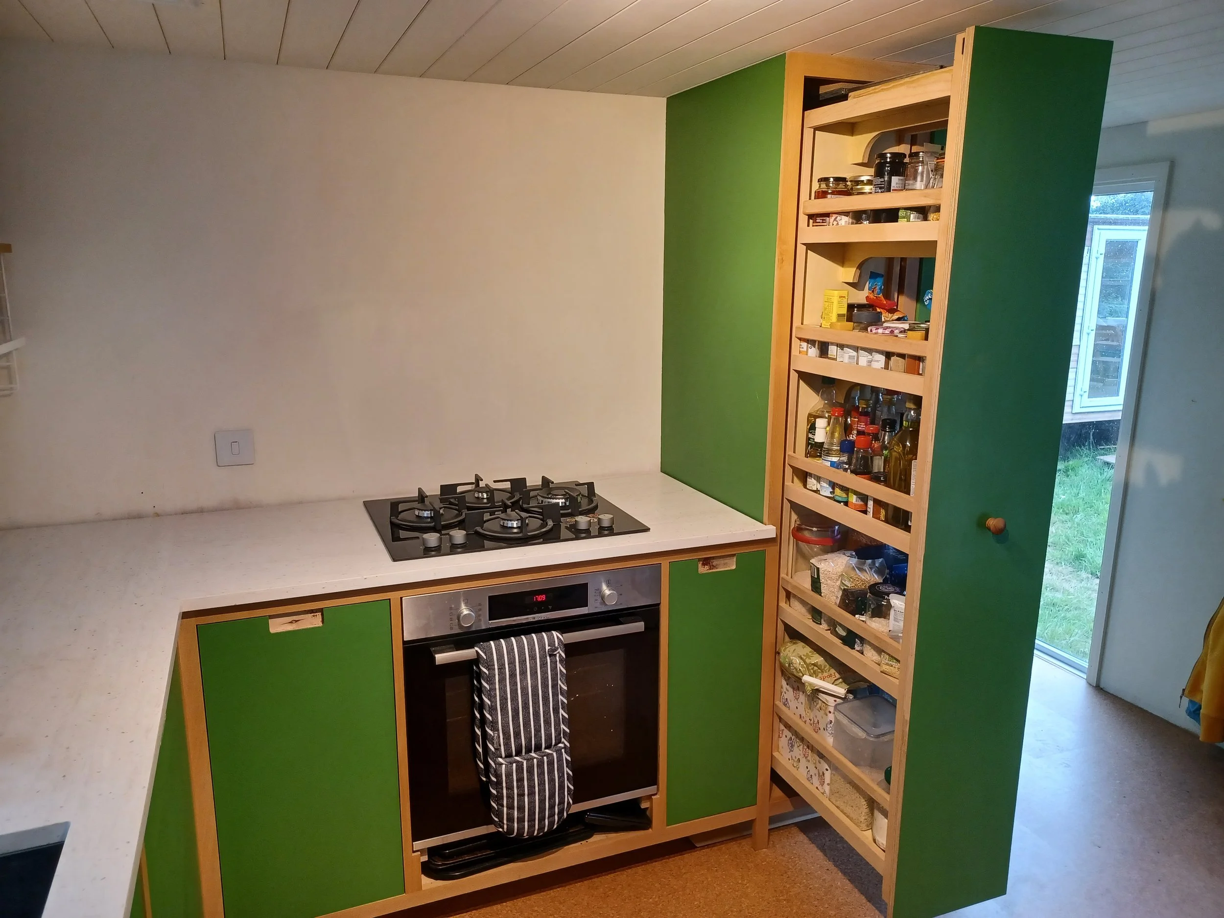 Kitchen corner with green cabinets, a built-in oven, a cooktop, and a wooden open pantry filled with spices and kitchen supplies. There is a window and a door leading outside.