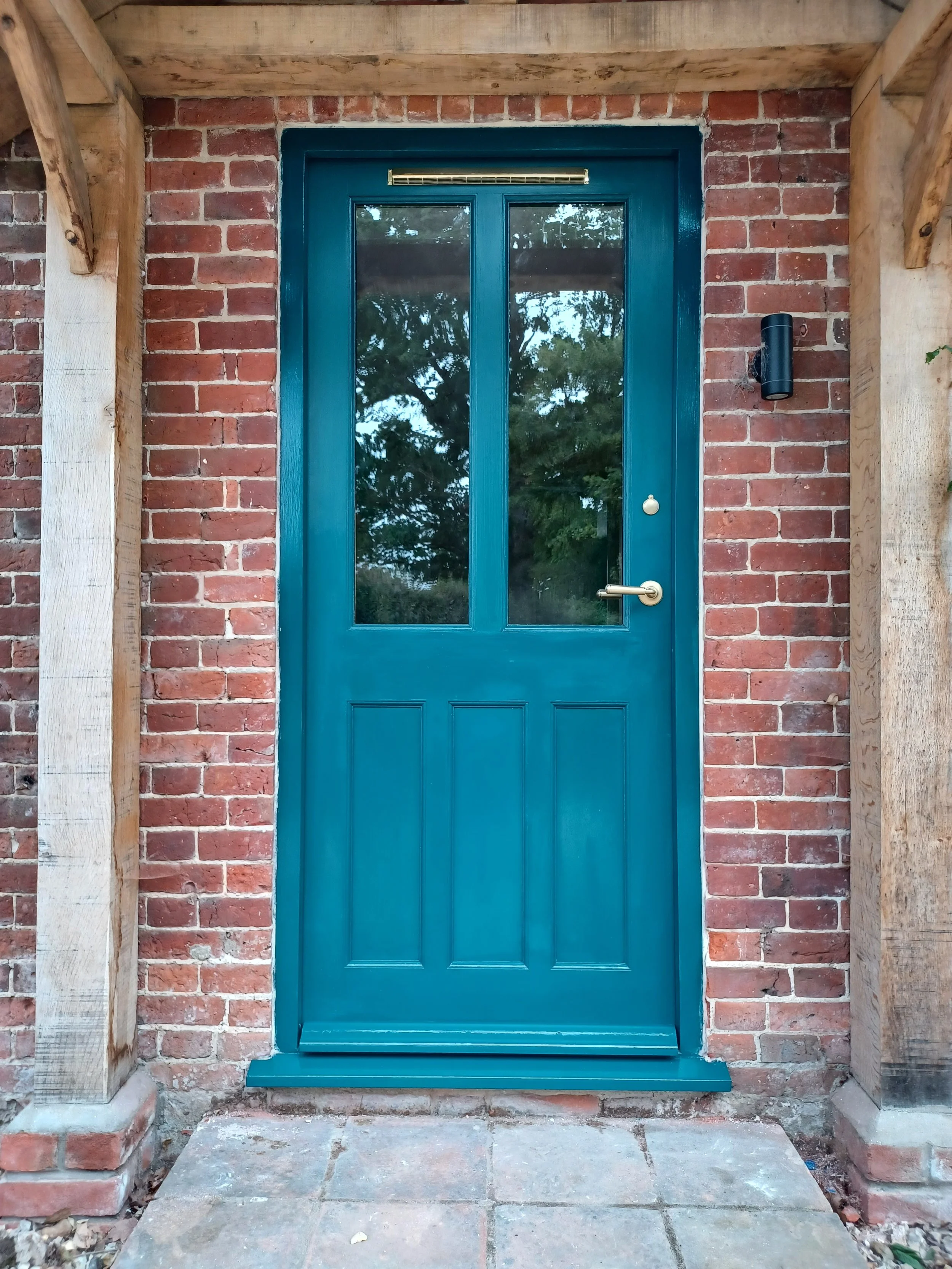 Blue front door with glass panels, surrounded by a brick wall and wooden framing, with a brick step and concrete patio in front.